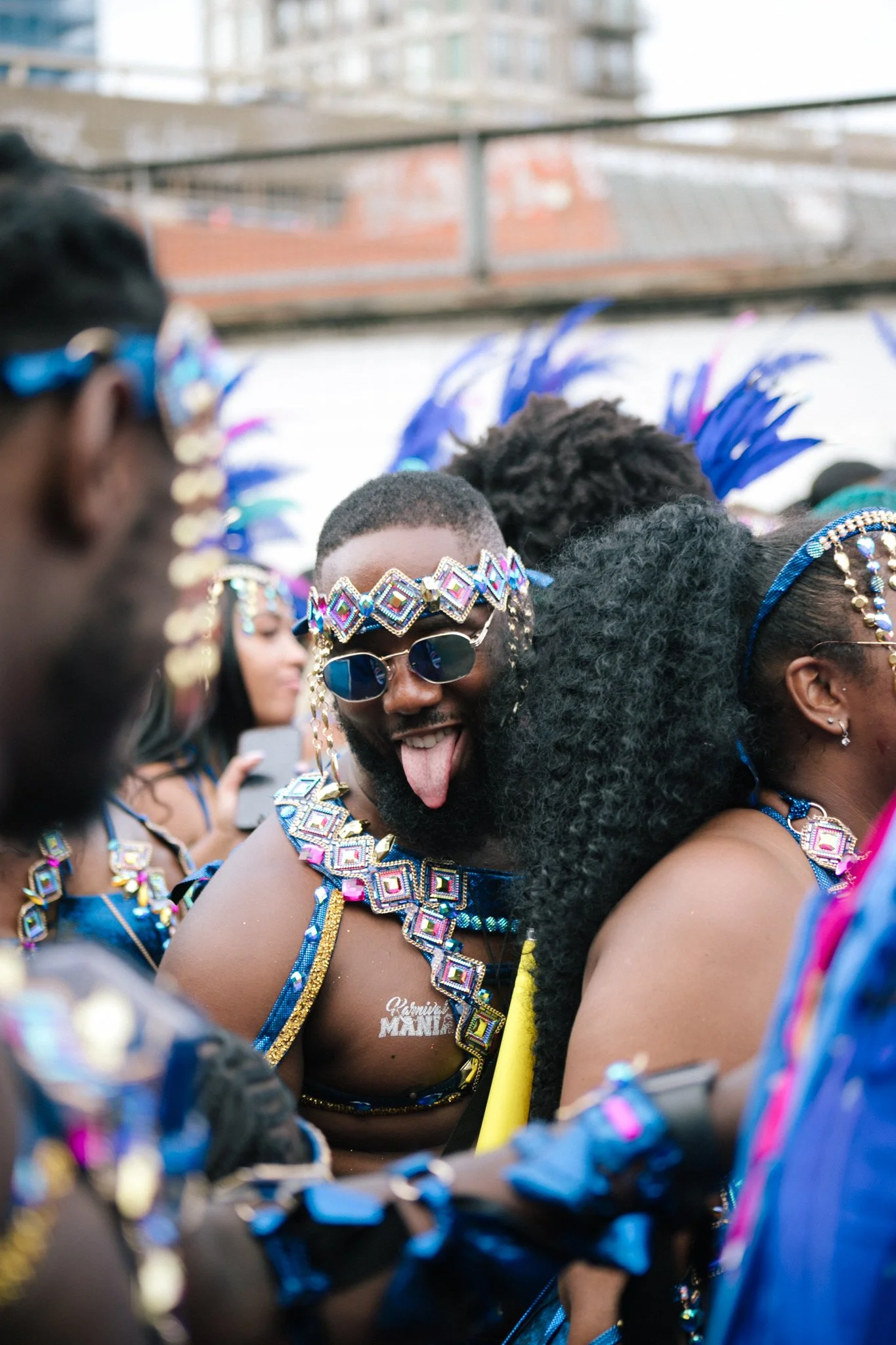 A group of people wearing colorful, elaborate costumes and accessories at a celebration or parade, with a focus on a man sticking out his tongue and smiling.