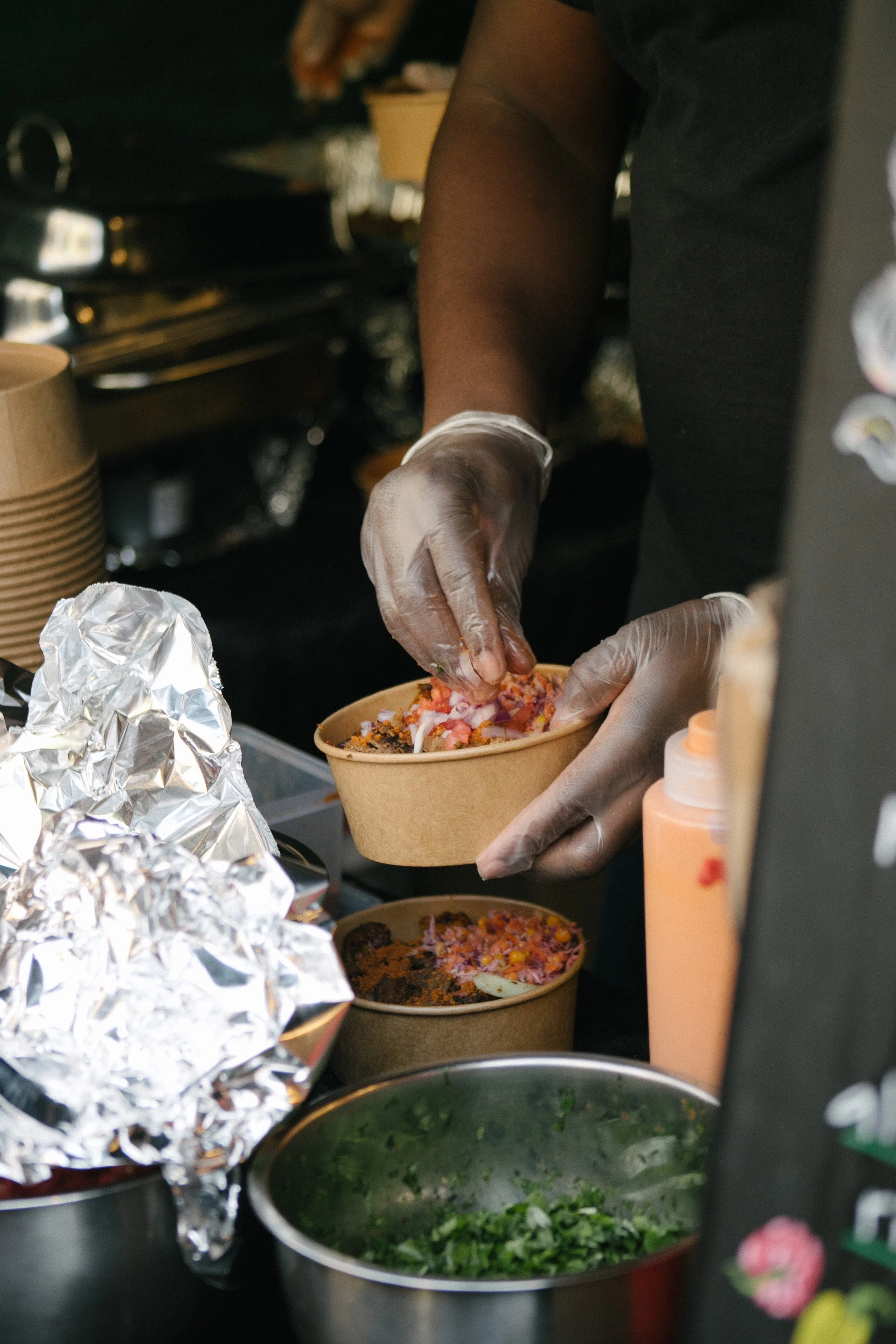 Person wearing gloves preparing food in a food stall, with bowls of chopped vegetables, salad, and food ingredients on the counter.
