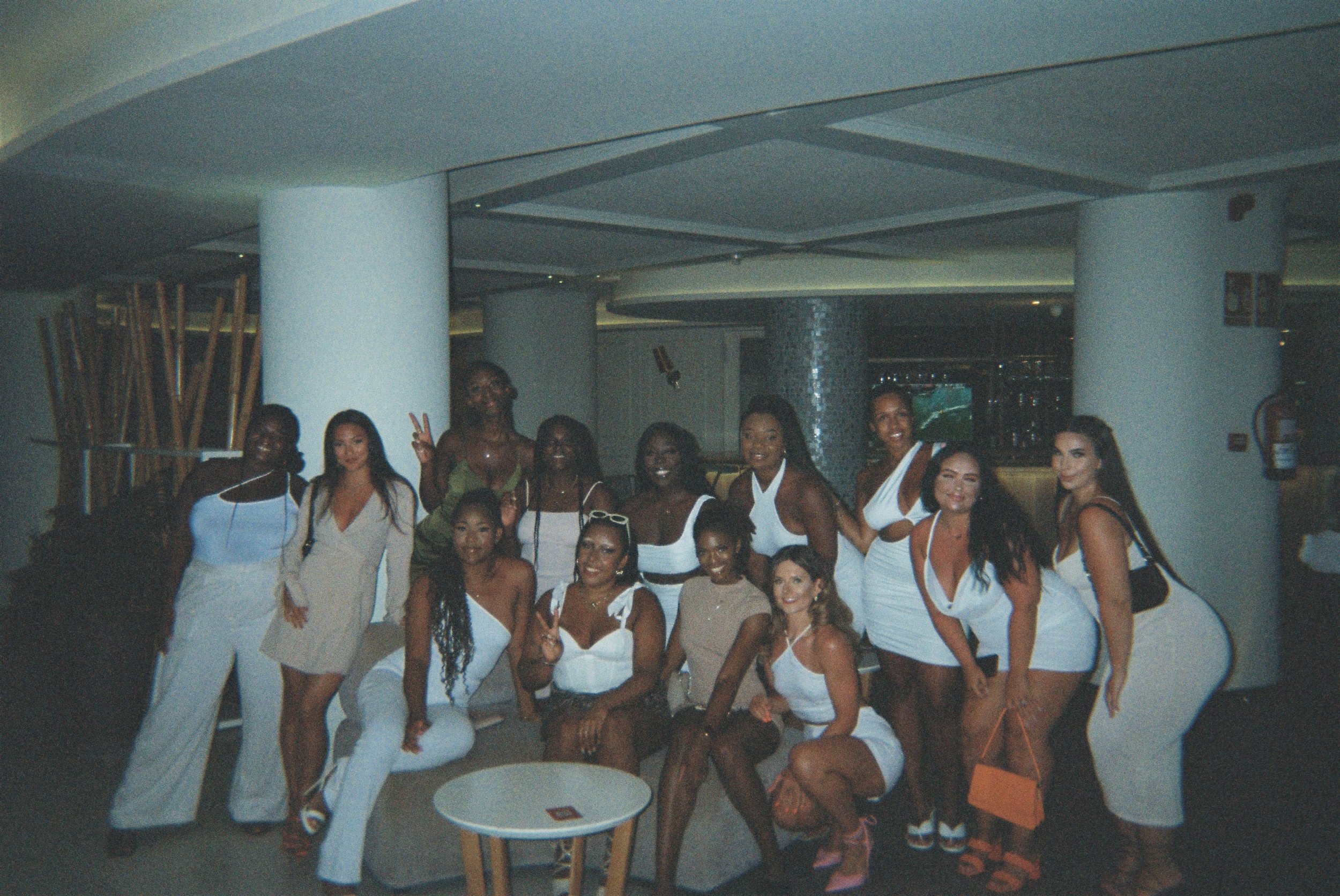 Group of women in white dresses posing together indoors, some making peace signs.