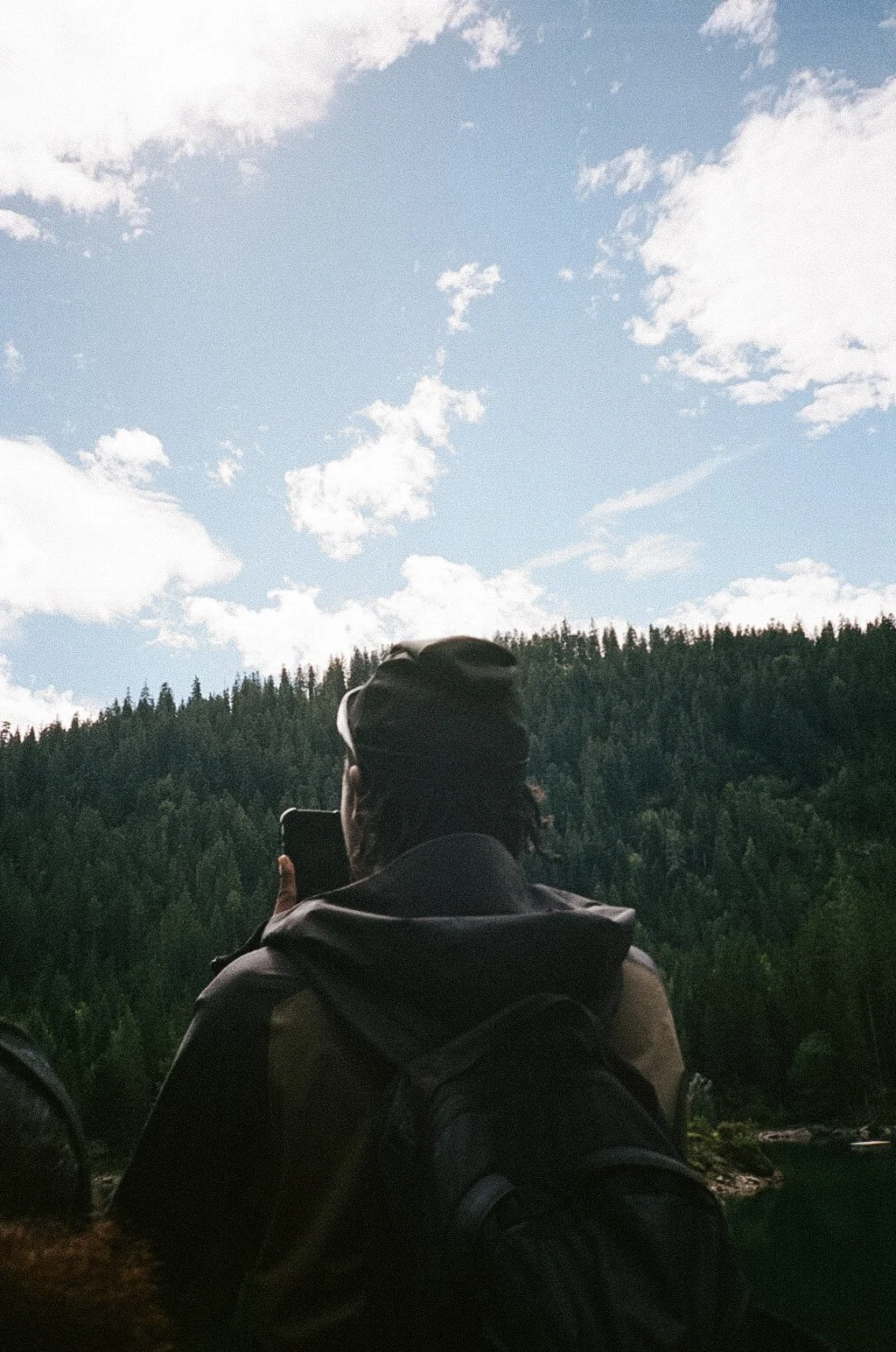Person with dark hair, wearing a dark jacket and a backpack, taking a photo with a smartphone of a forested mountain landscape under a partly cloudy sky.