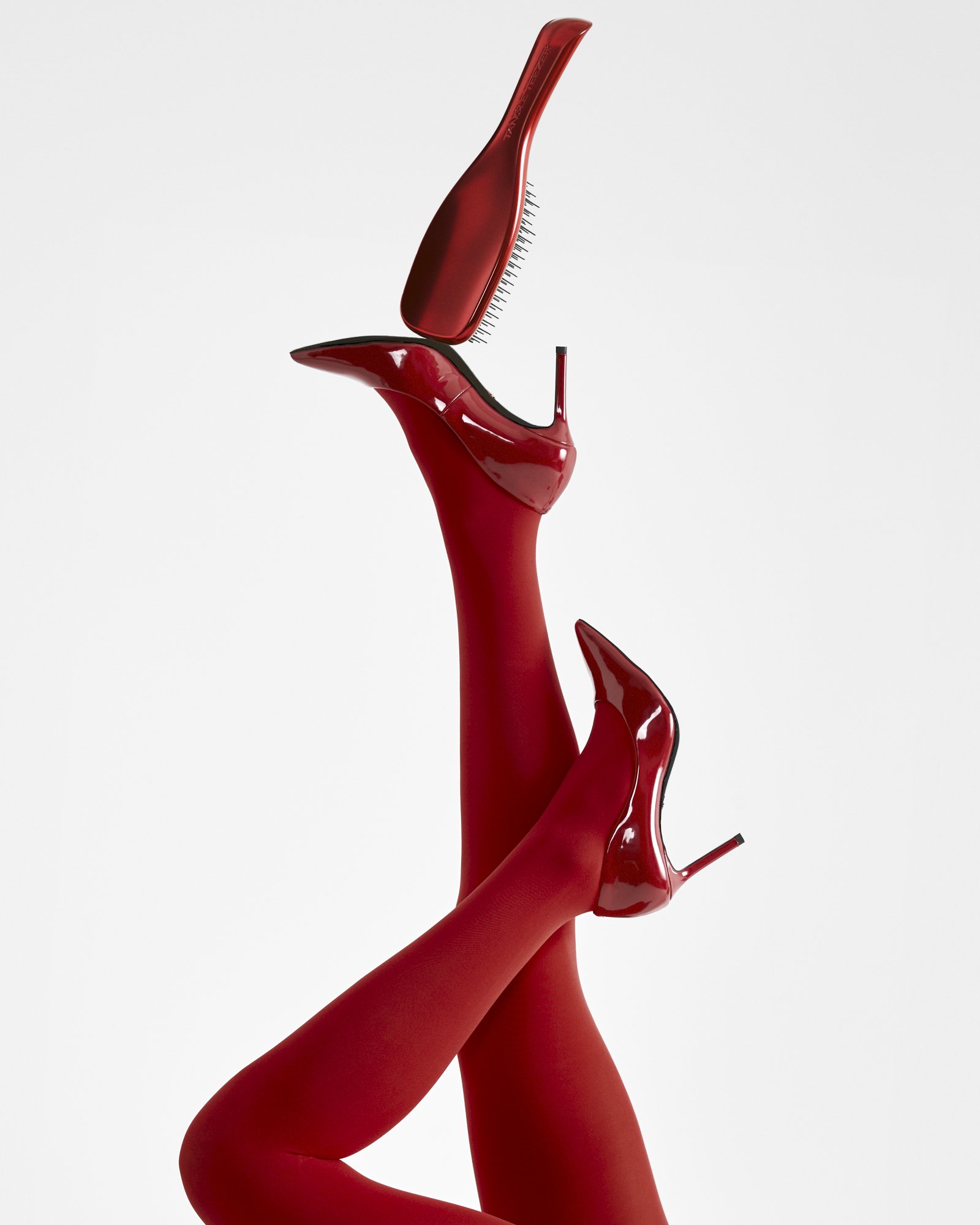 A red high-heeled shoe and a red hairbrush with a curved handle are arranged to mimic a pair of legs and a foot, set against a white background.