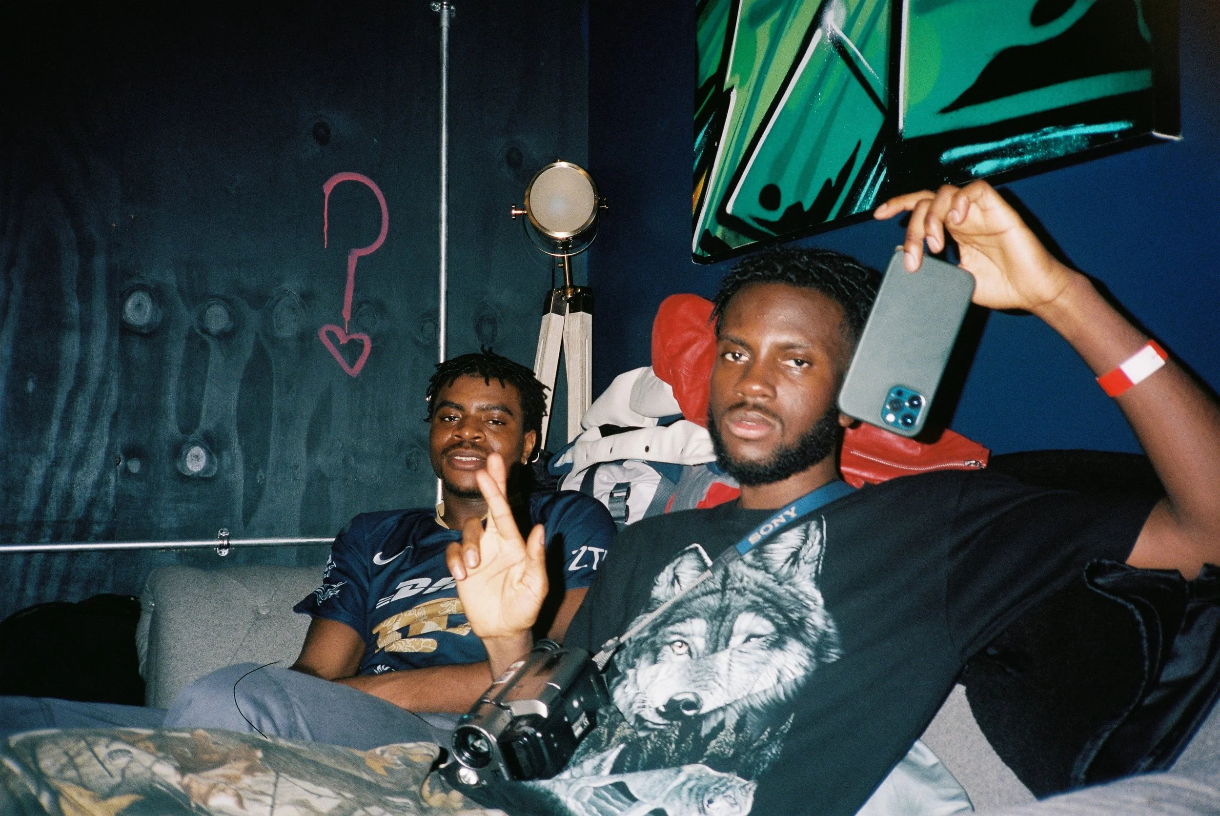 Two young men sitting on a couch in a room with dark walls, one taking a selfie with a smartphone, and the other making a peace sign. There is a pink question mark with a heart on the wall behind them, and a colorful abstract artwork on the wall to t