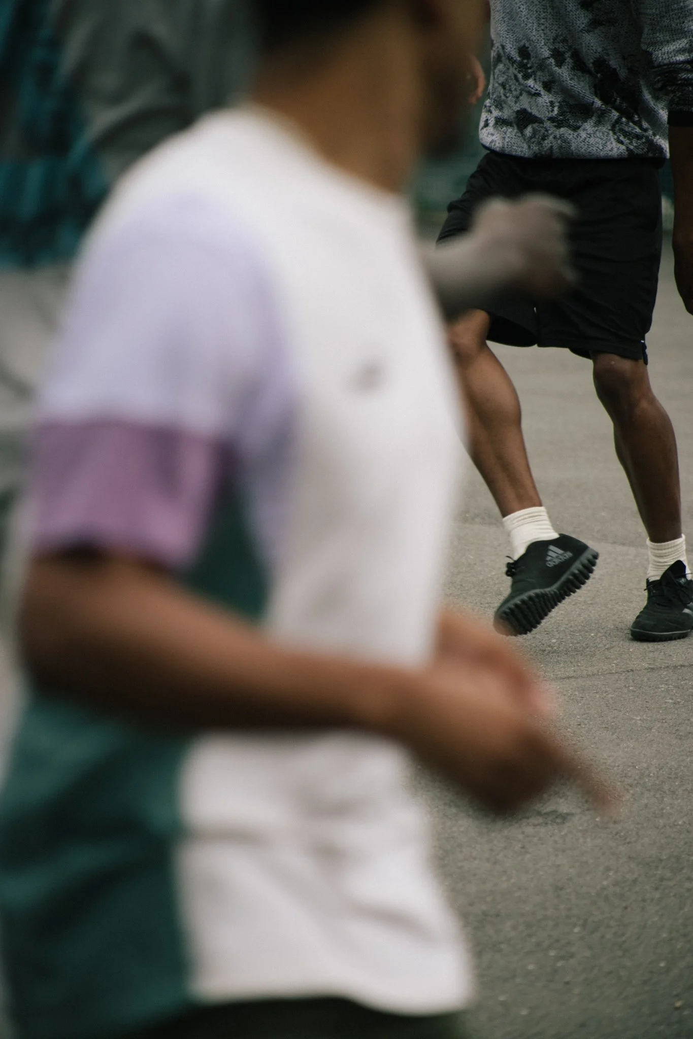 Blurred image of young men playing football outdoors, with one man in the foreground wearing a white and purple shirt and another in the background wearing a gray or patterned shirt and black shorts.