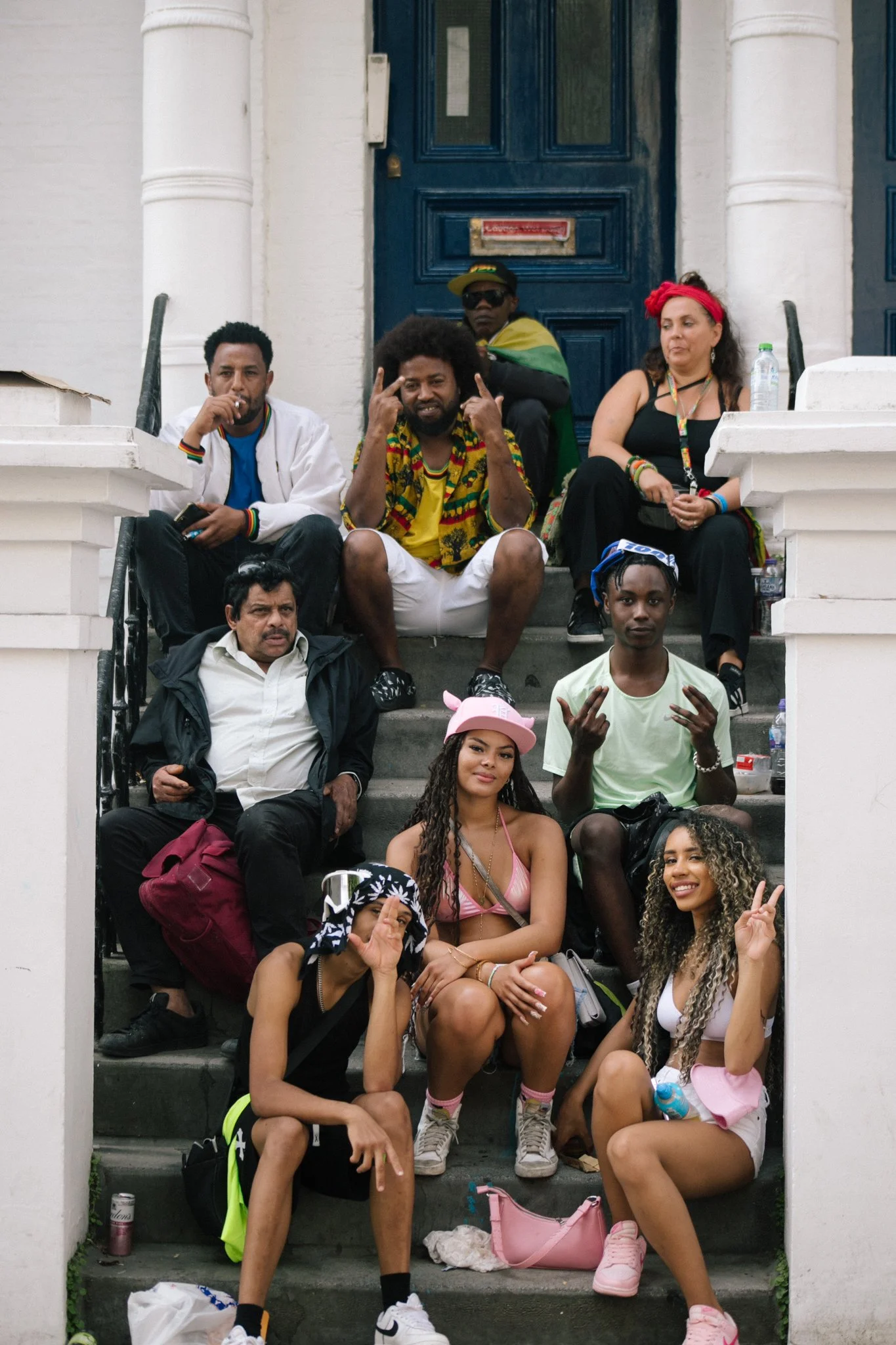 Group of people sitting on the stairs outside a building, some wearing casual attire with colorful accessories, making peace signs, and posing for the photo.