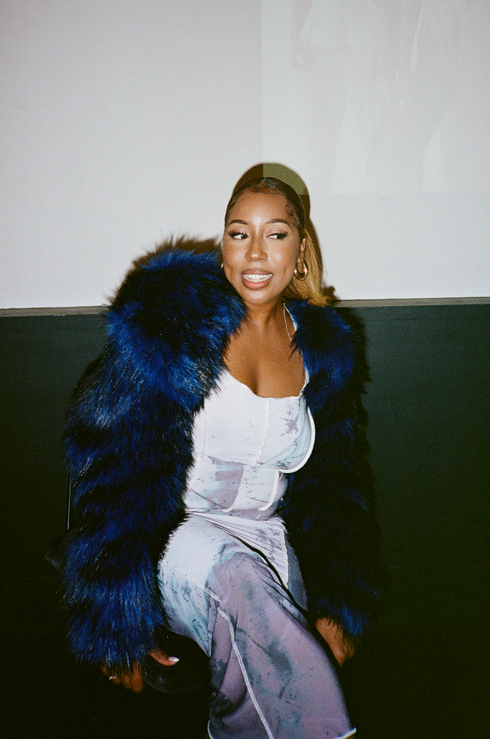 A woman wearing a blue and black fur coat and a light purple tie-dye co-ord, sitting against a white and black wall.