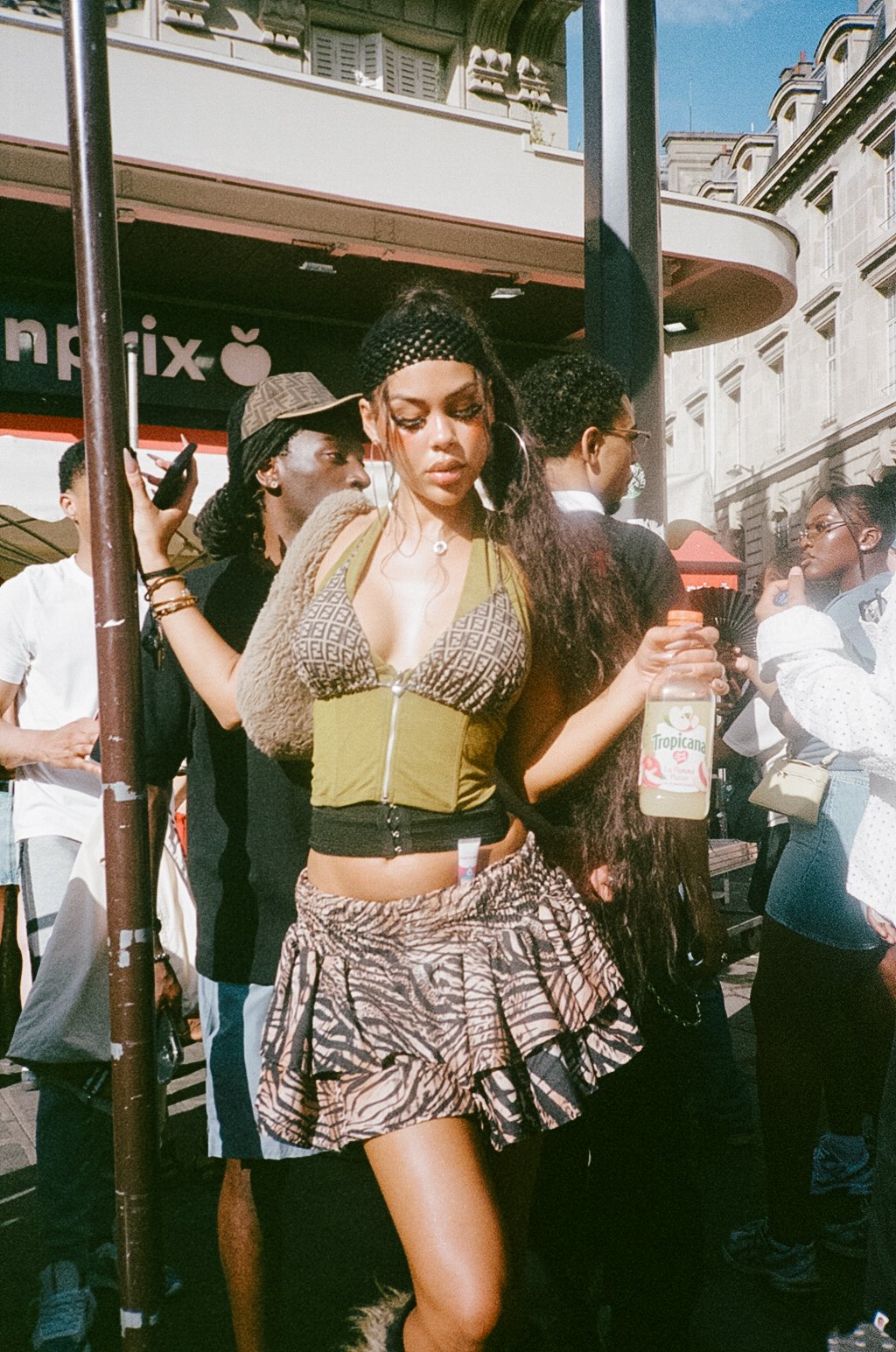 A young woman with long dark hair, wearing a fashionable outfit with a patterned crop top, a ruffled skirt, and accessories, standing outdoors on a city street with other people around her and a storefront in the background.