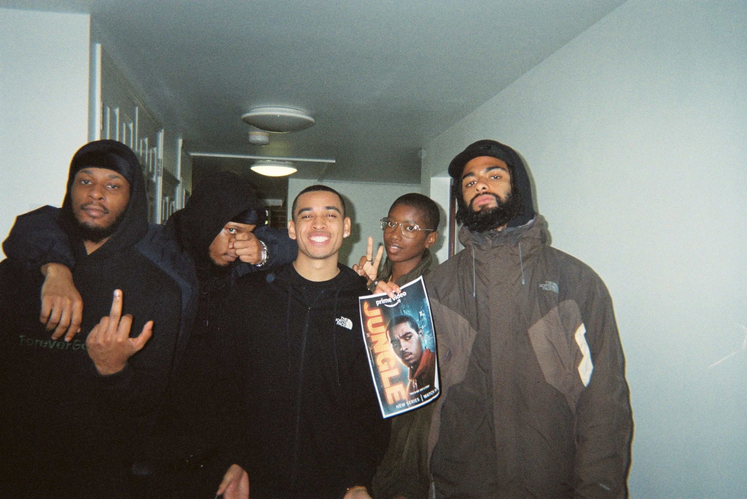 Group of four young men and one woman posing indoors, one holding a poster, some making hand gestures, wearing casual jackets and hoodies.