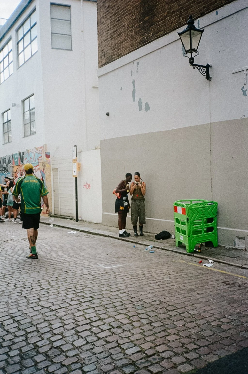 Street scene with two women looking at a smartphone, a man walking away, a group of people in the background, and a bright green plastic barrier on the sidewalk.