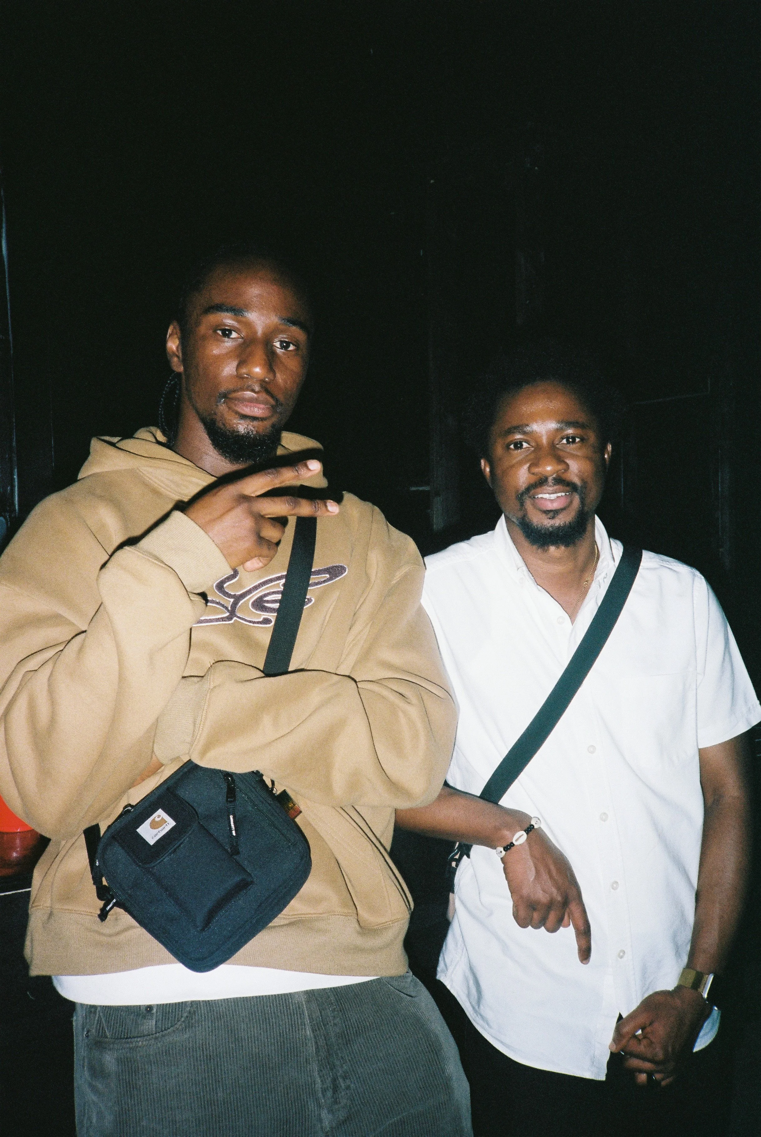 Two men standing together in a dark background, one making a peace sign, both smiling and looking at the camera.