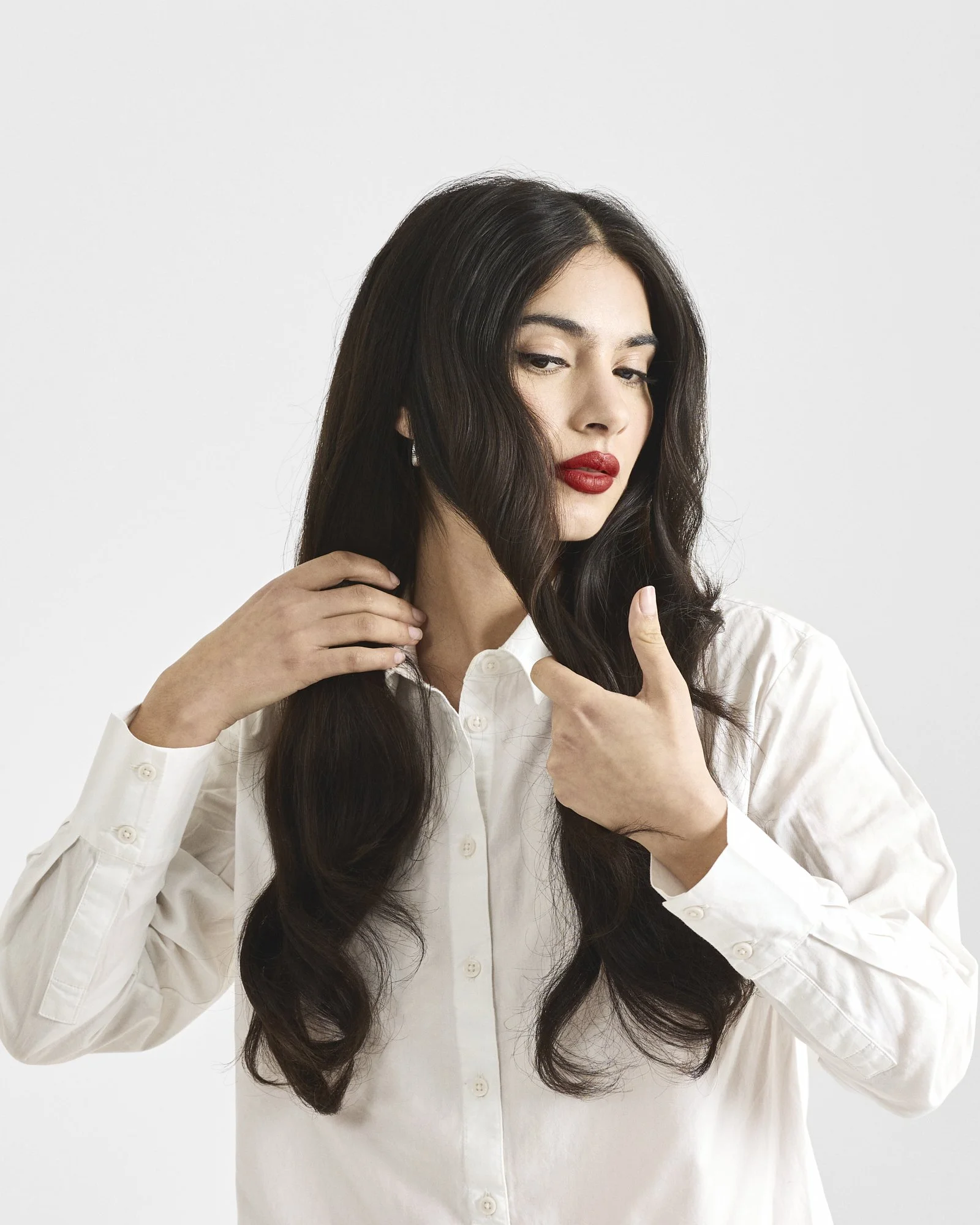 A woman with dark, wavy hair and red lipstick wearing a white shirt, looking away from camera, separating her hair - against a plain light background.