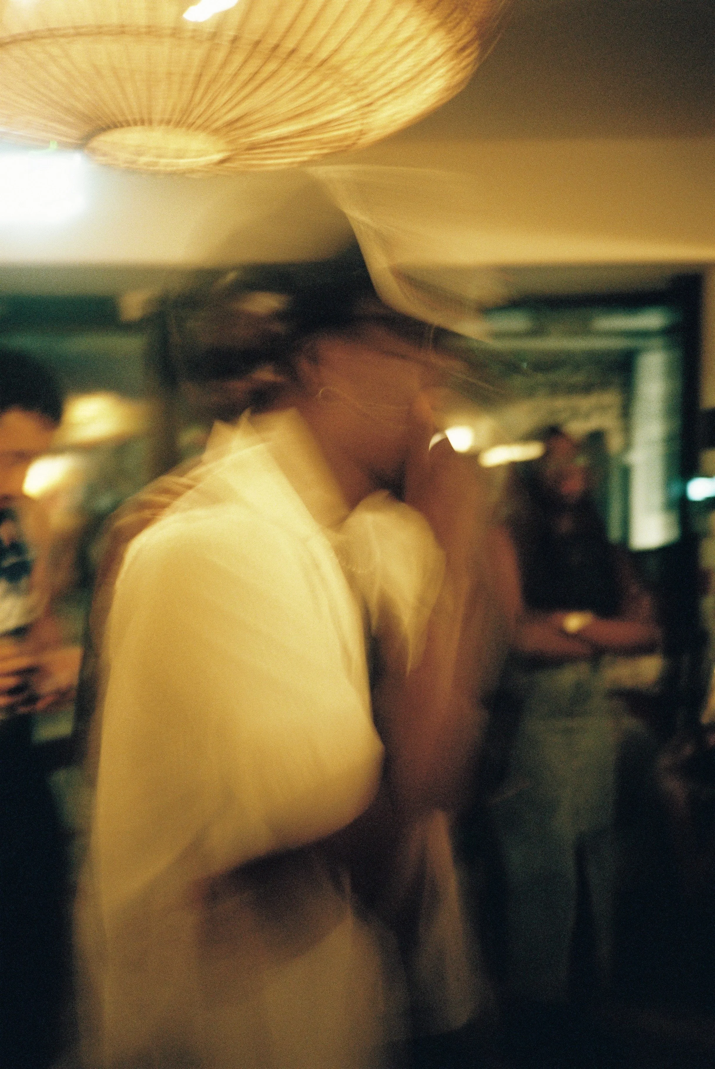 A man blurred in the foreground of a dimly lit room with a large woven pendant light hanging from the ceiling. There are other people in the background, some with drinking glasses.