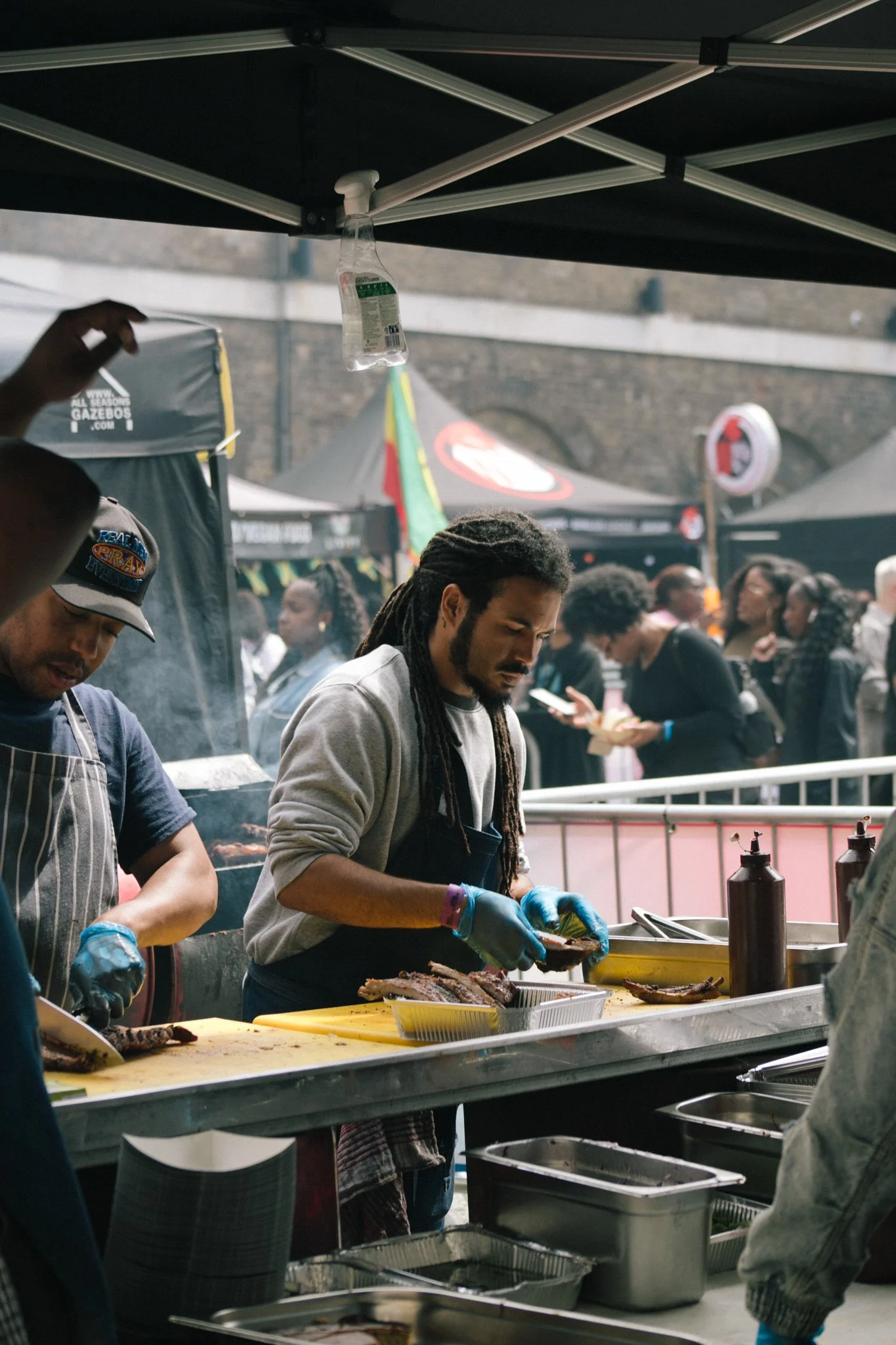 People preparing food, particularly jerk chicken, at an outdoor food stall with tents in the background.