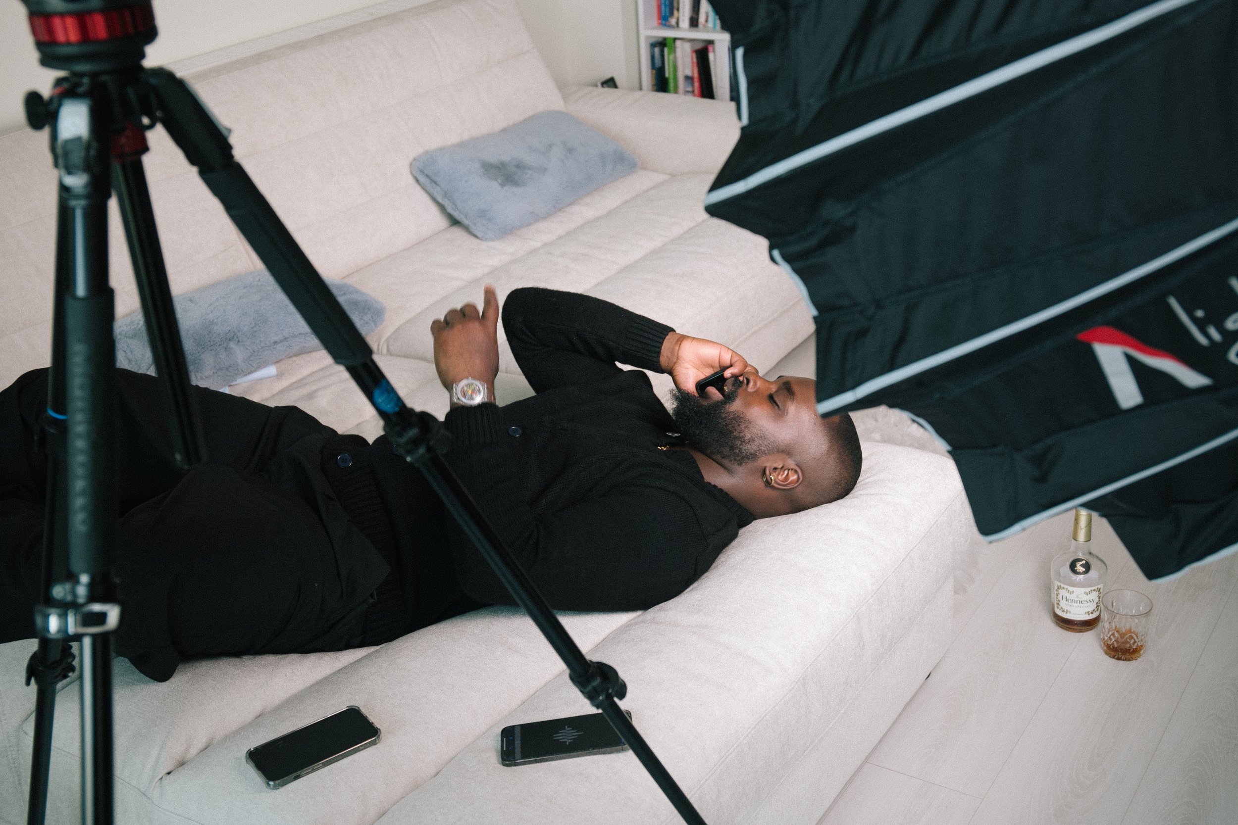 A man is lying on a white couch, holding a cellphone in his right hand, which rests on his face. On the floor there are two smartphones and a glass of Hennessy brand cognac. Part of a camera setup or lighting equipment is visible in the foreground.
