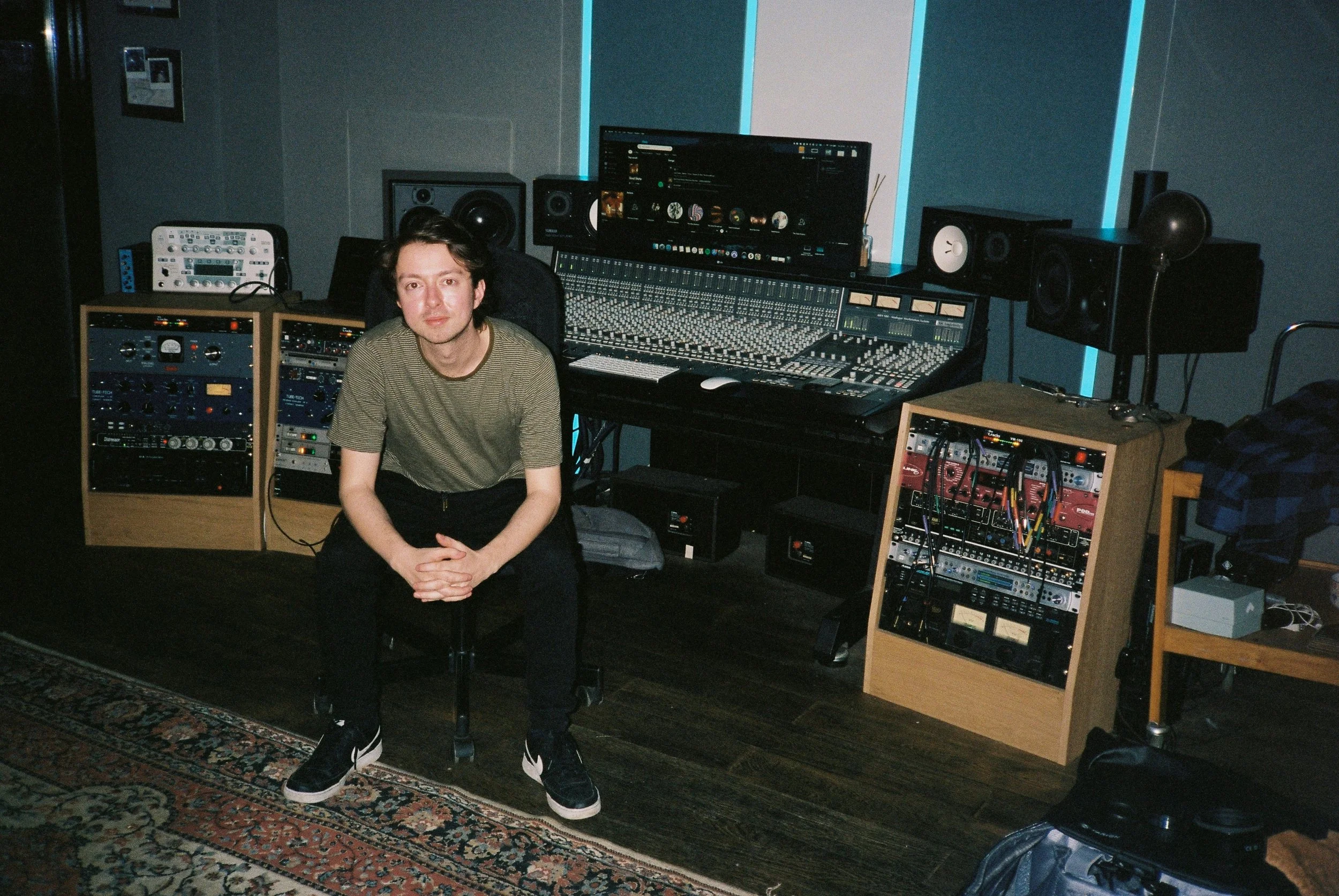 A young man sits in a recording studio surrounded by audio equipment, including a mixing console, speakers, and synthesizers. He is wearing a striped t-shirt and black sneakers, sitting on a chair on a patterned rug.