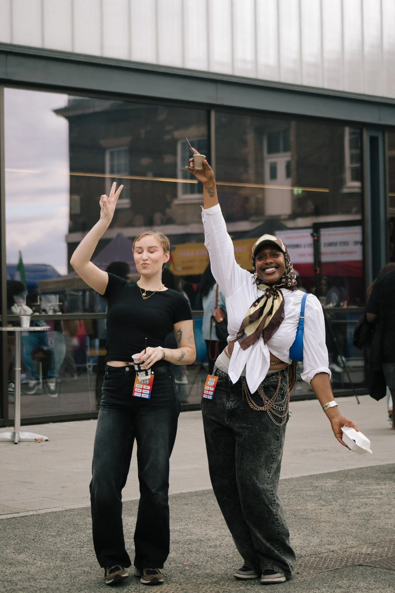 Two young women standing outdoors in front of a glass building, smiling with their hands in the air and making peace signs. Both have women festival/event badges hanging around their necks.