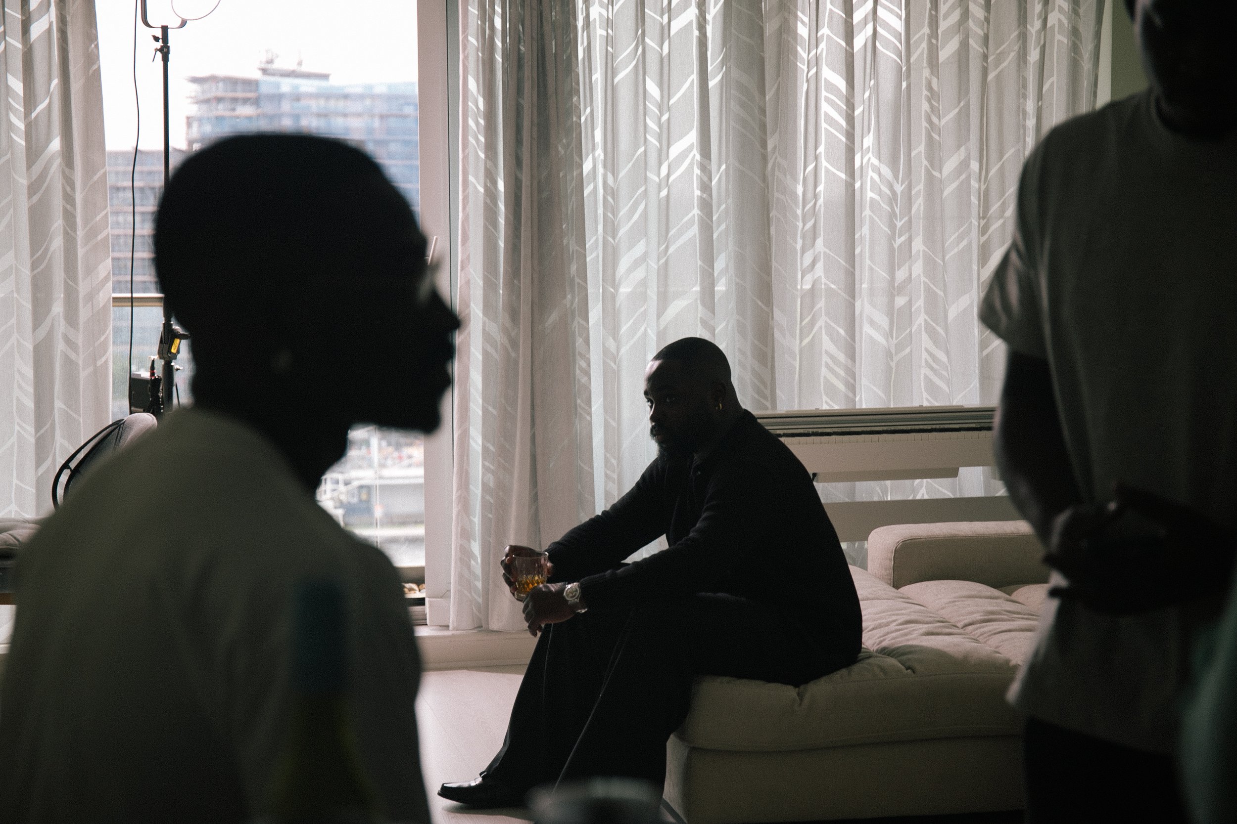 Silhouettes of four people in a room with sheer patterned curtains, a window, and a cityscape outside. One person is sitting on a bed holding a drink, while others are standing or sitting, engaged in conversation.