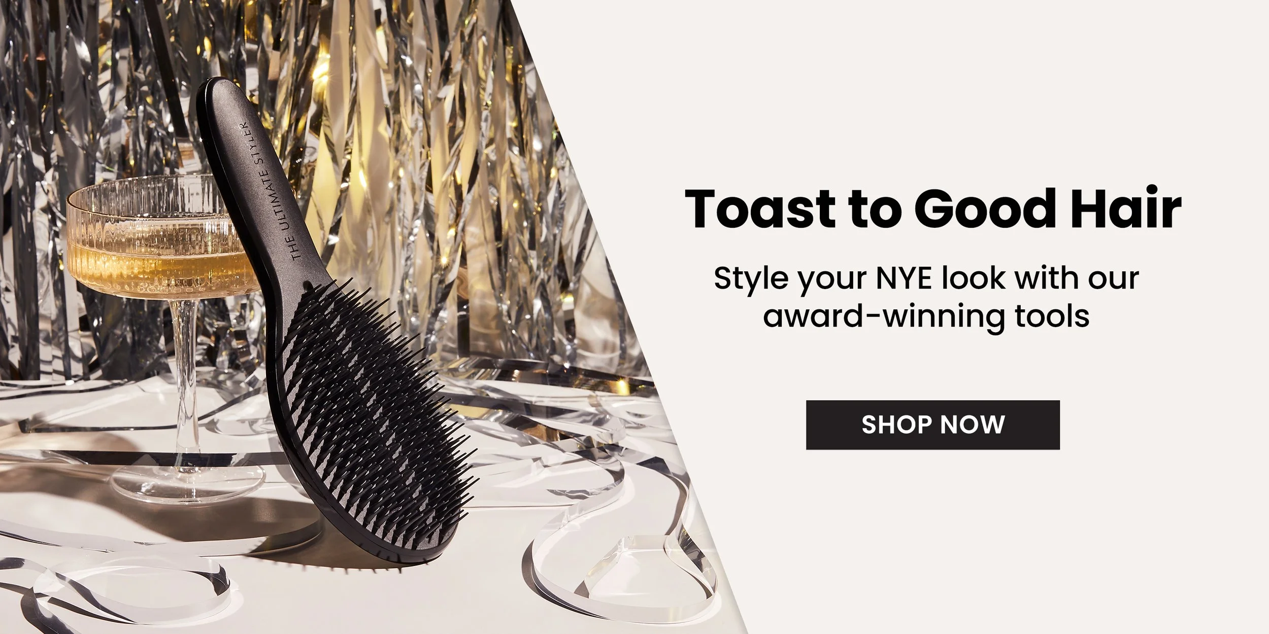 A hairbrush with black bristles and a handle labeled 'The Ultimate Styler' sitting on a surface. Next to it, there is a champagne coupe glass filled with a golden beverage, with silver and gold metallic streamers and fairy lights in the background.