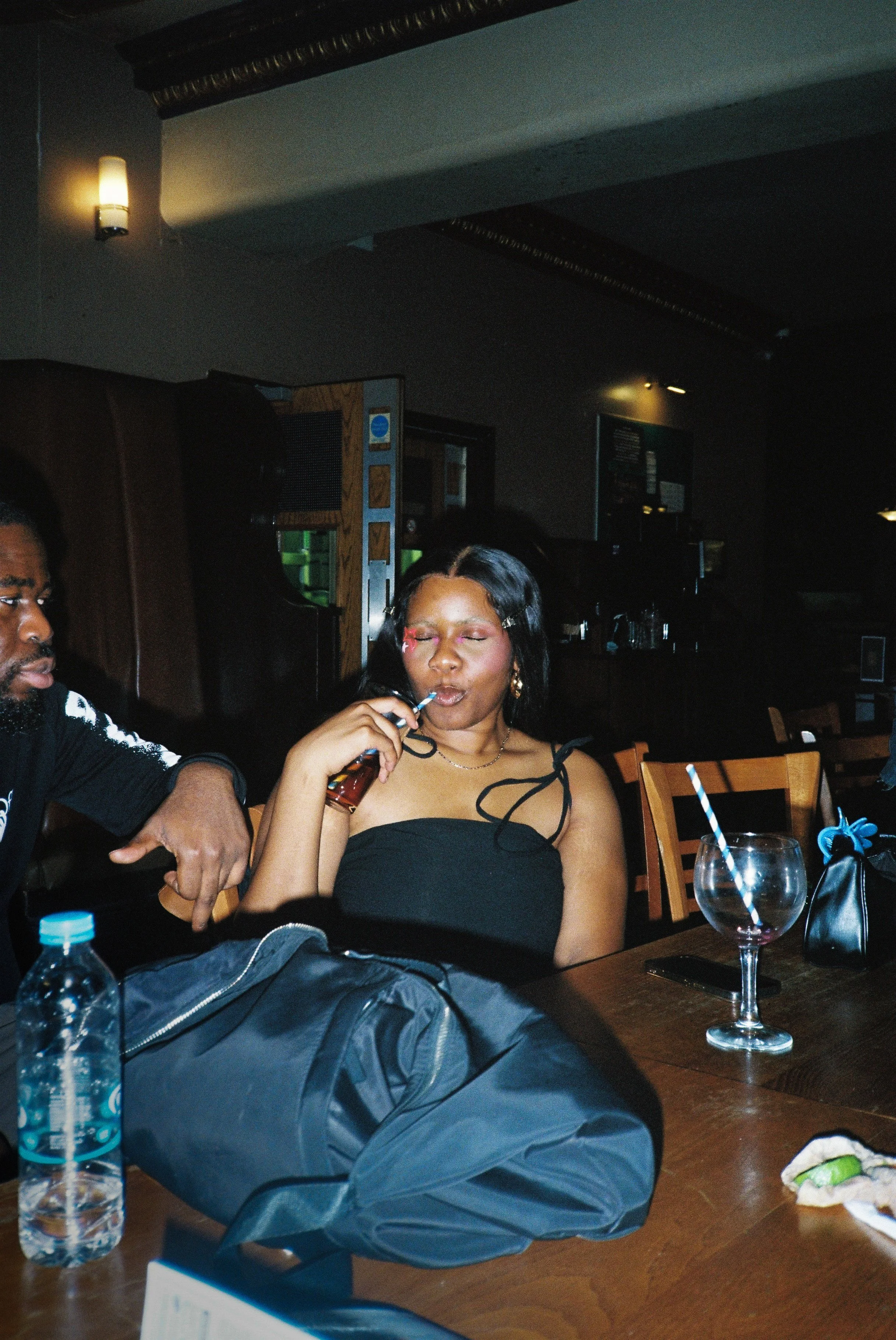 A woman with closed eyes sitting at a dinner table in a pub, with a man sitting beside her and various items on the table including water bottles, a glass with a straw, and a partially eaten sandwich.