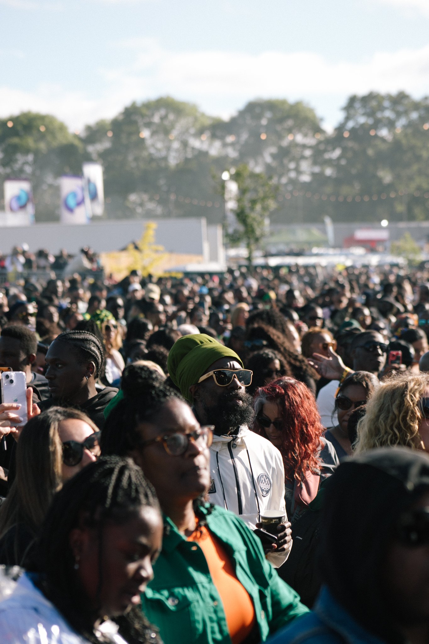 A large crowd of people gathered outdoors, many wearing sunglasses and colorful clothing, with some taking photos. In the background, there are trees, flags, and string lights, indicating a festive or event setting during daytime.