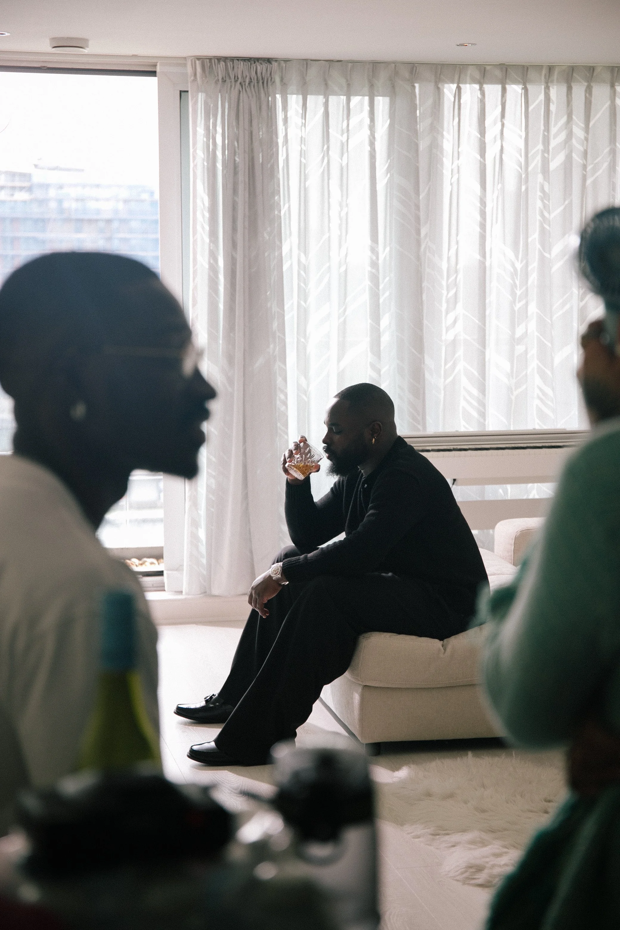 Man wearing all black sitting on a beige sofa, drinking from a glass, with a large window with white curtains in the background. Two people are partially visible in the foreground.