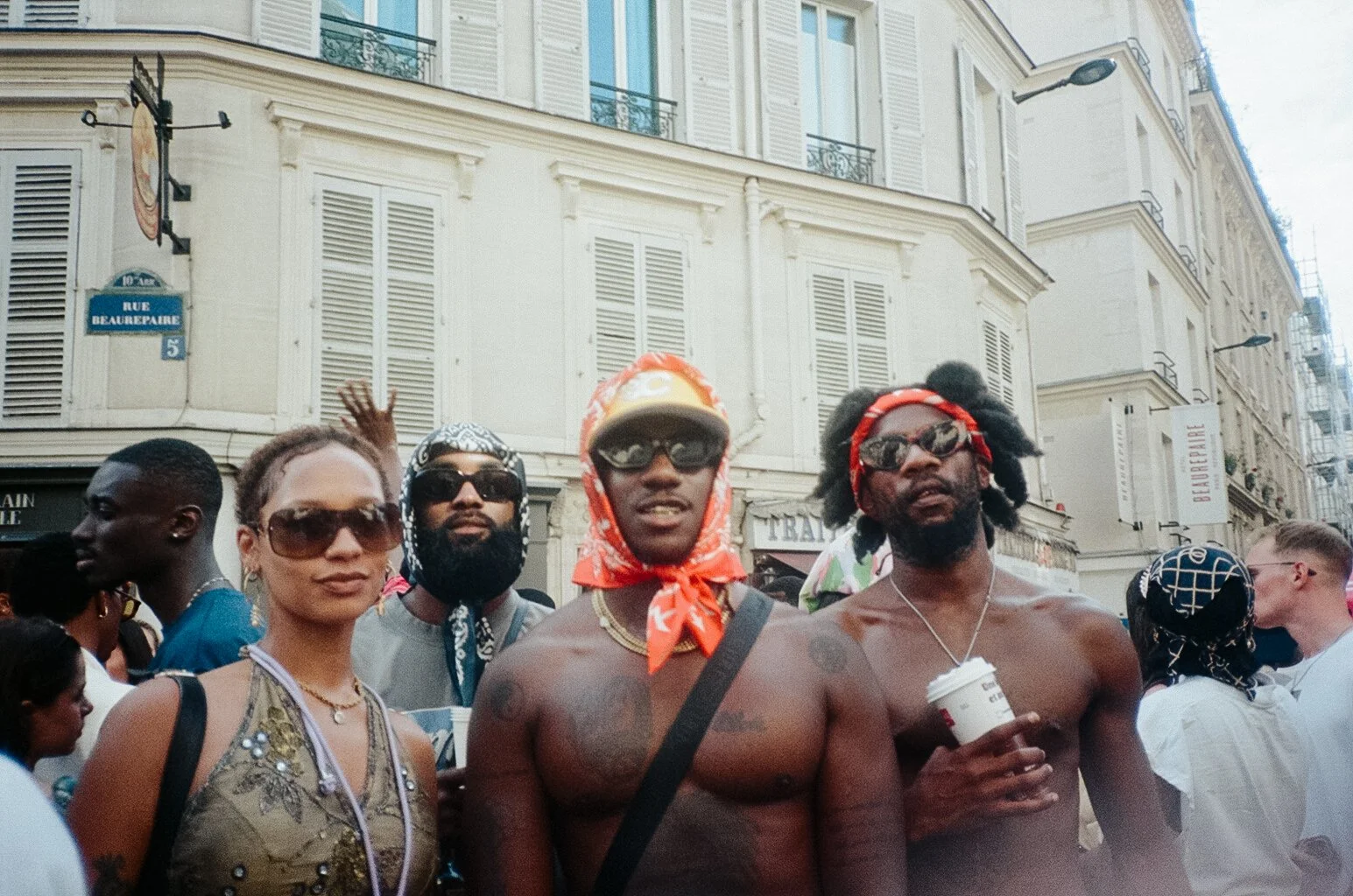 A diverse group of people gathered outdoors on a street with Parisian-style buildings in the background. Some are shirtless, wearing sunglasses and bandanas, and others are dressed casually, holding drinks. The scene suggests a festive atmosphere.
