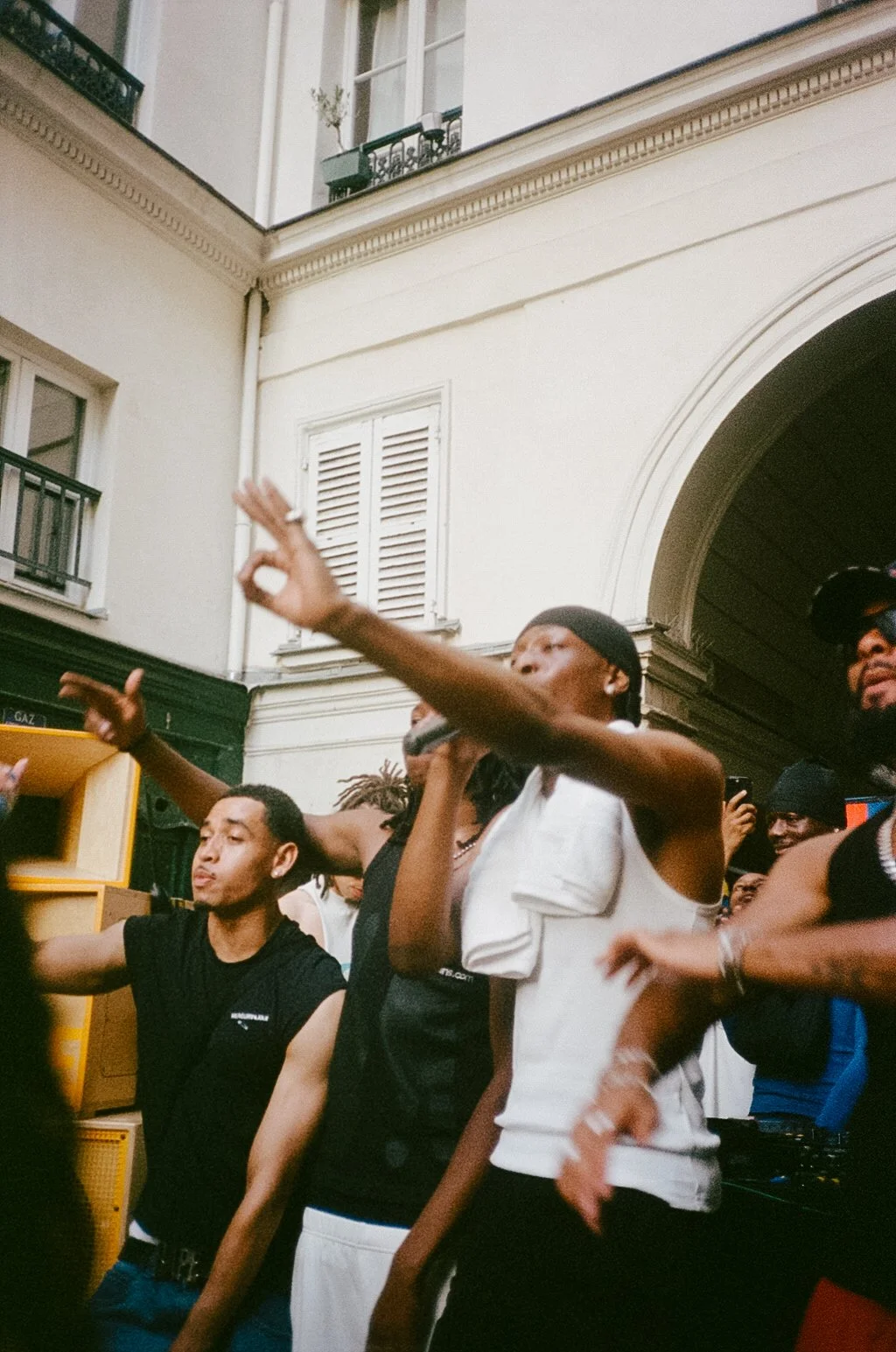 A group of people are performing outdoors, with one person in the center holding a microphone and gesturing with their hand. Others are around, some with arms raised or holding phones, in a lively and engaged atmosphere.