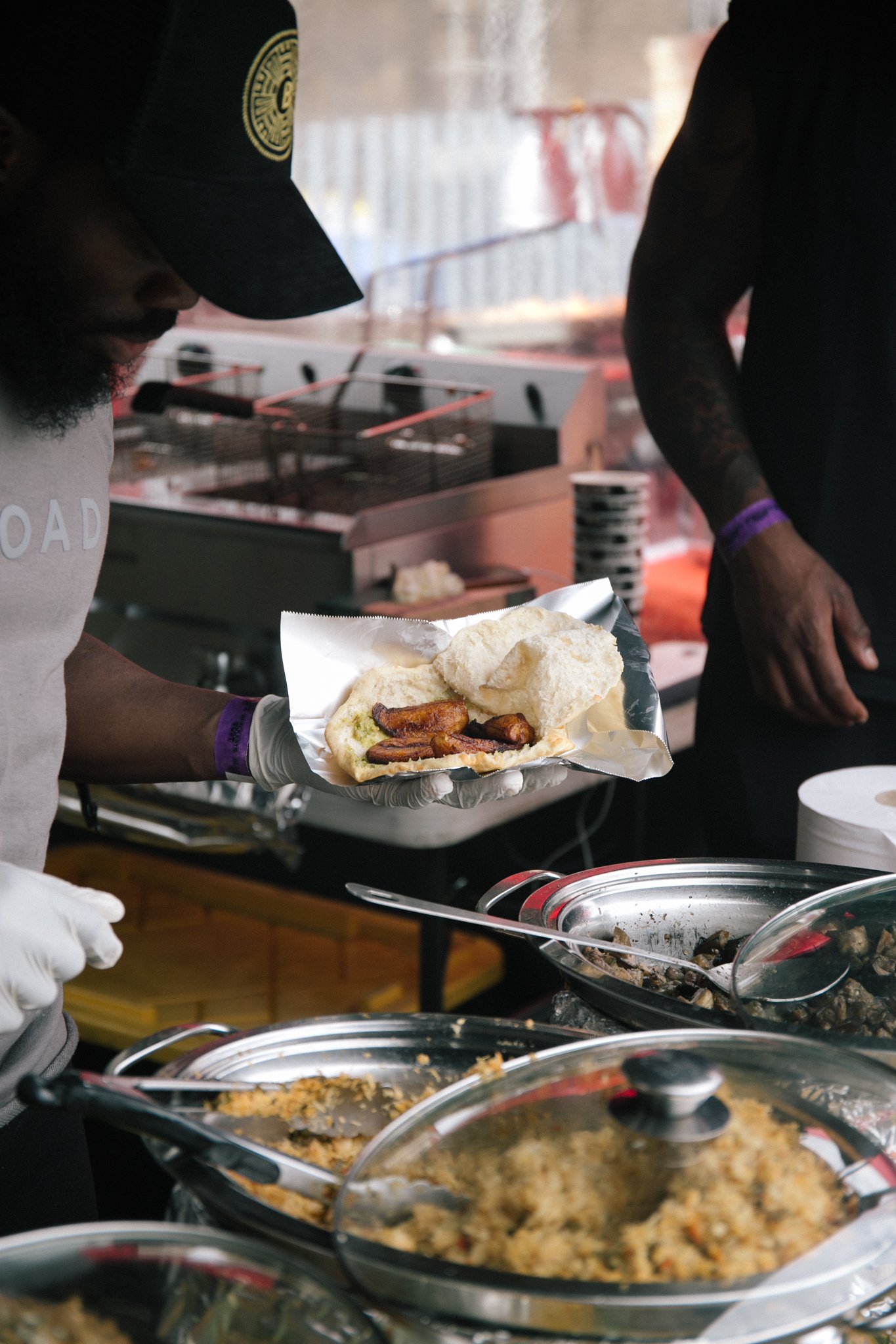 Person holding an open bokit (a Guadeloupean fried bread sandwhich) with fried plantain inside at a food counter, with others serving food in the background.