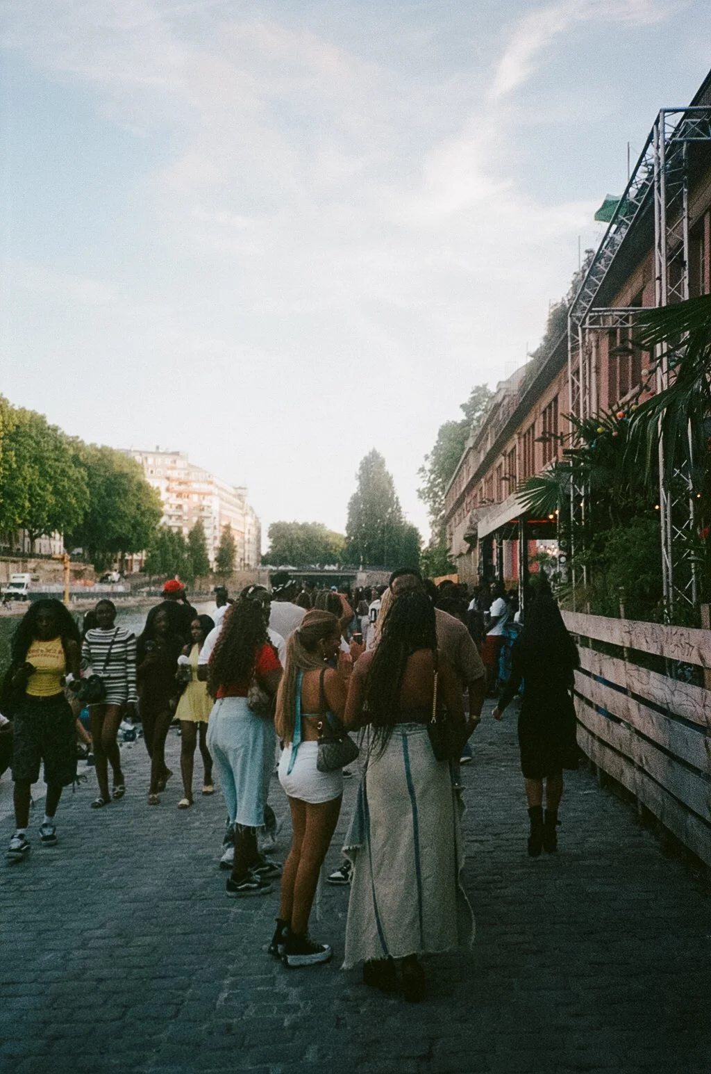 People gathered outdoors on a cobblestone street near a river, with buildings and trees in the background, and a structure on the right side of the image.