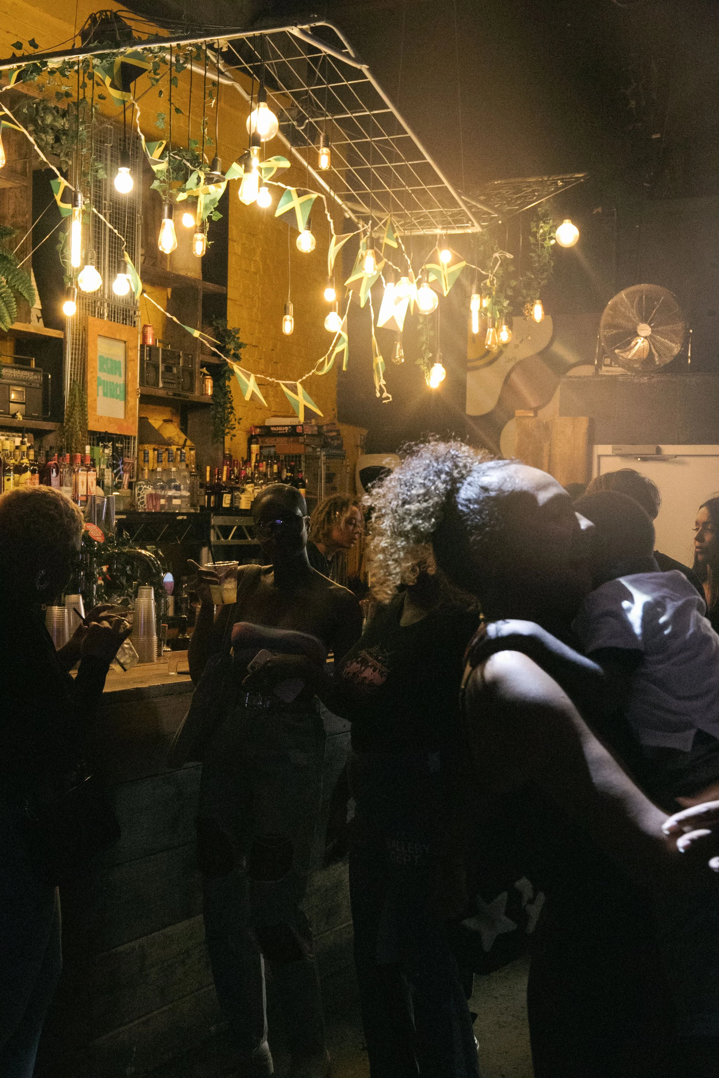A group of people gathering at a bar with warm lighting. The bar has a yellow brick wall with shelves of alcohol bottles and a stereo system. Decor includes string lights, hanging bulbs, and green and yellow flags. The scene appears to be in a lively