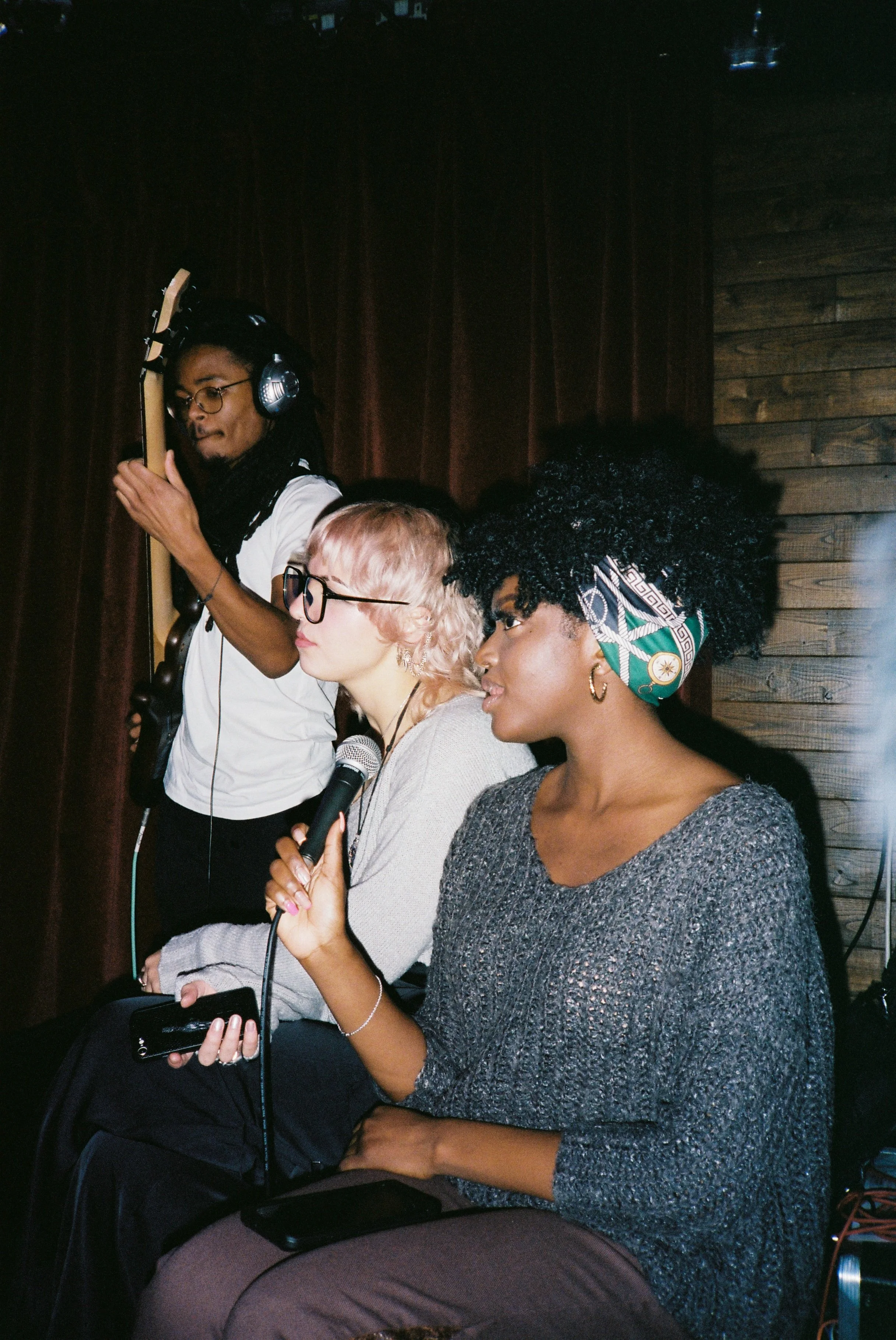 Two women sitting in a row, holding microphones, with a man standing with a guitar behind them. The women singing or speaking, in a dimly lit indoor setting with wooden wall panels.