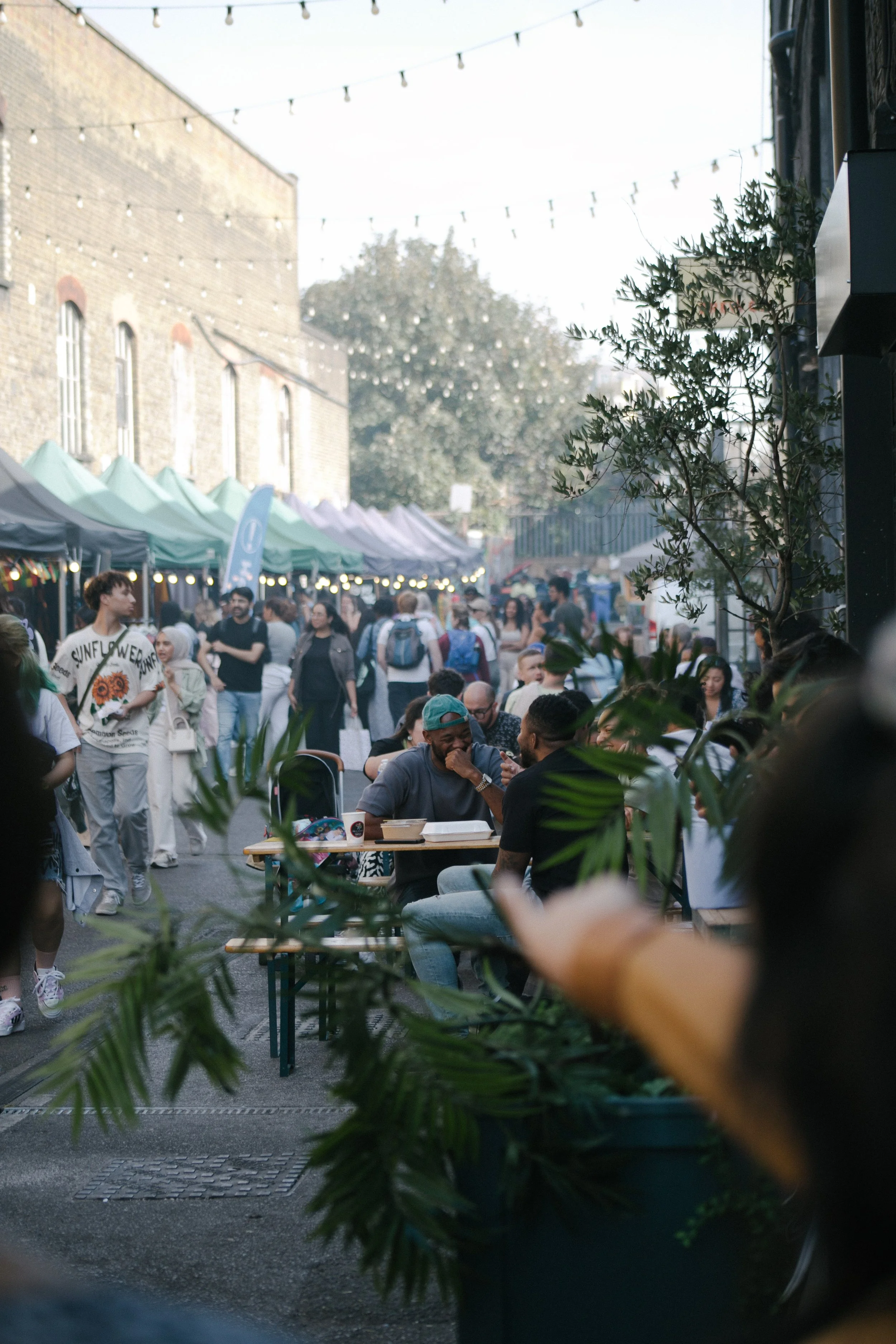 A crowded outdoor market or street fair with tents, string lights, and people walking and sitting at tables. Some trees and buildings are visible in the background.