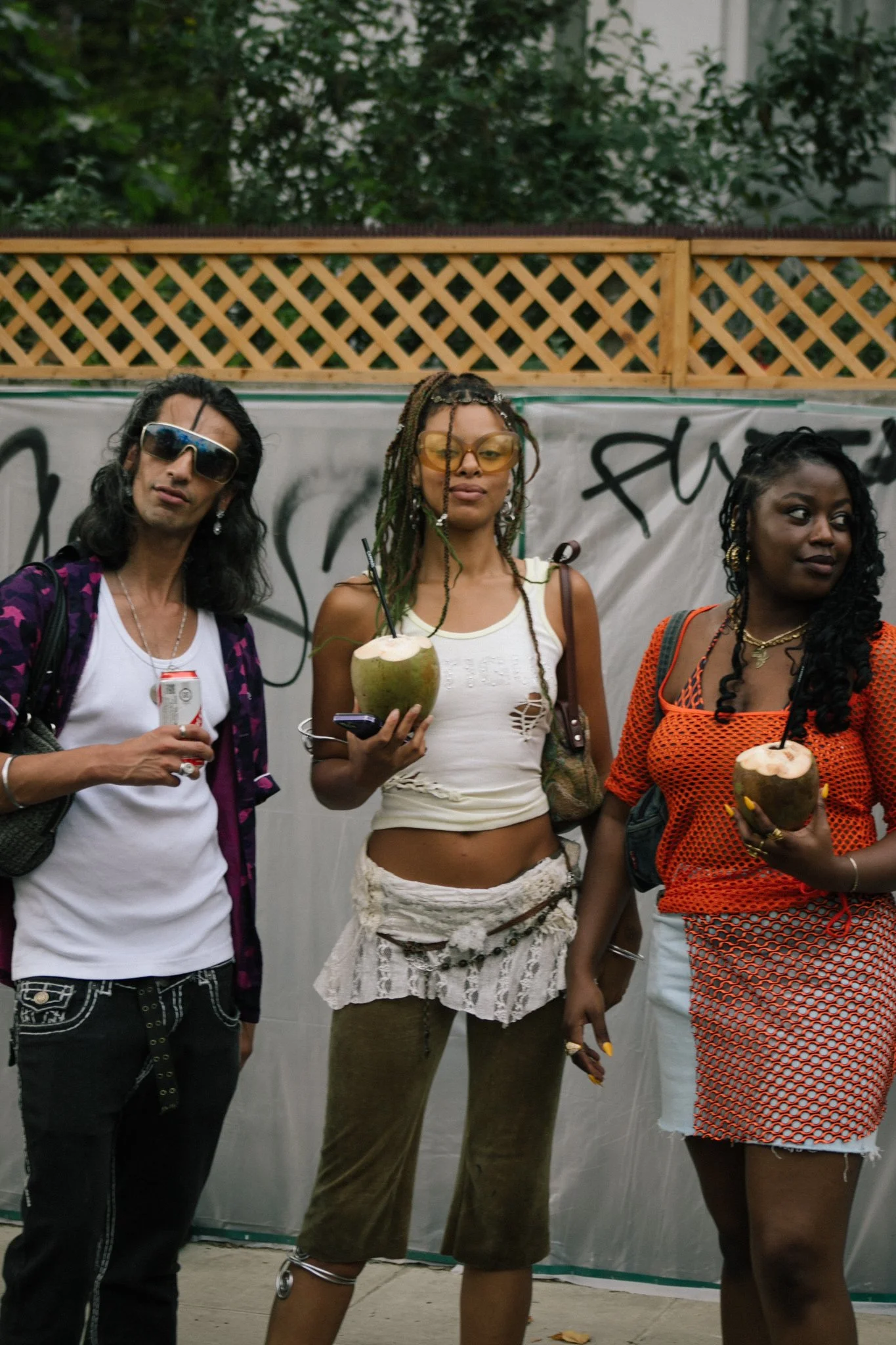 Three people standing together outdoors, each holding a coconut with a straw, near a graffiti wall and a wooden fence in the background.