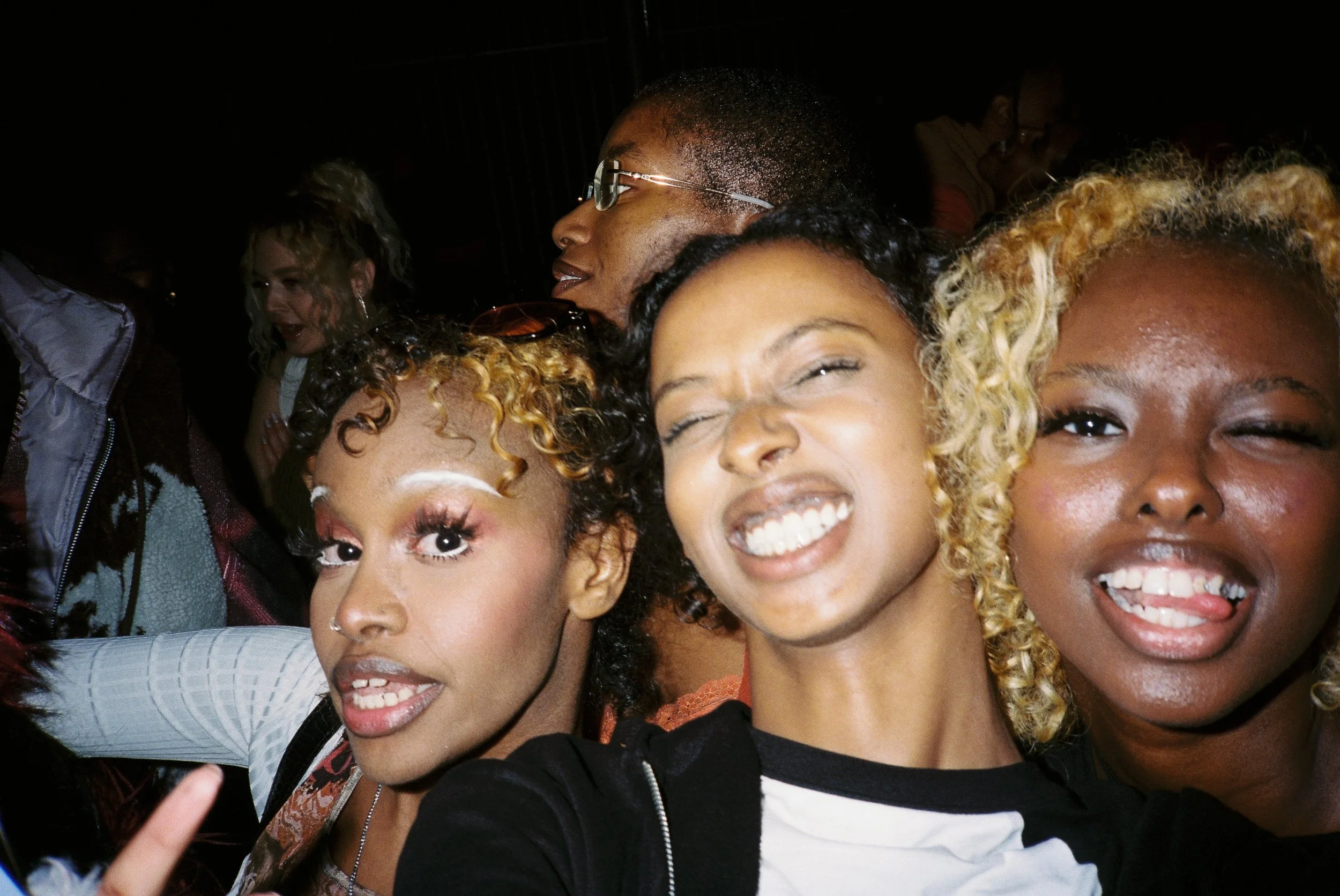 Group of women taking a close-up selfie, smiling, in dim lighting.