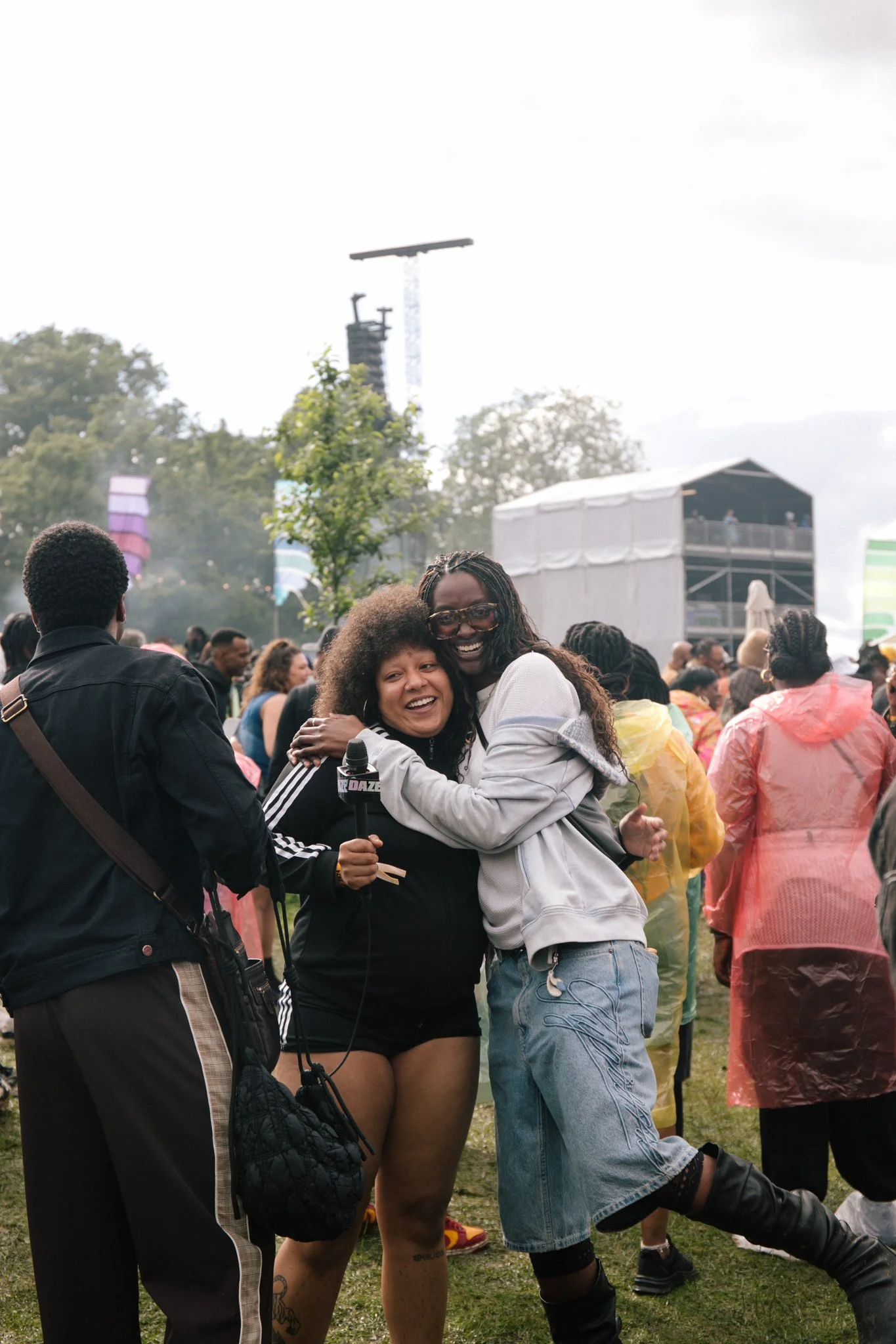 People gathered outdoors at an event, smiling, hugging, and enjoying the day. In the foreground, two women are hugging, one holding a microphone. Others are wearing rain ponchos, and a stage and trees are visible in the background.