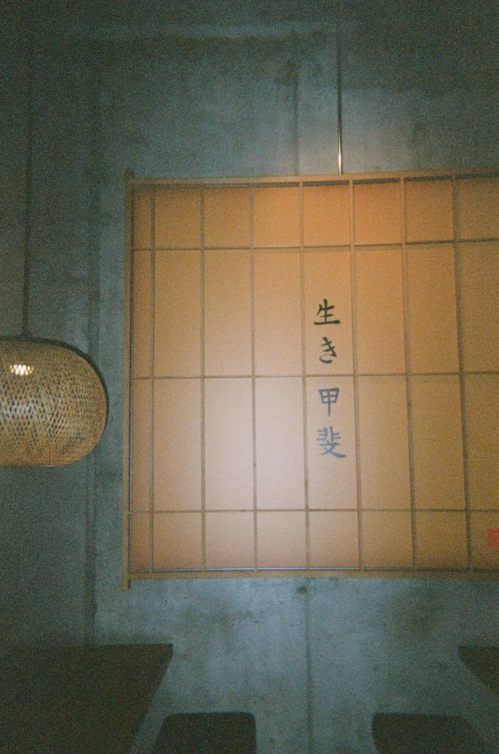 A Japanese signboard with Japanese characters written vertically, placed on a tiled wall. There is a wicker lantern on the left side of the image.