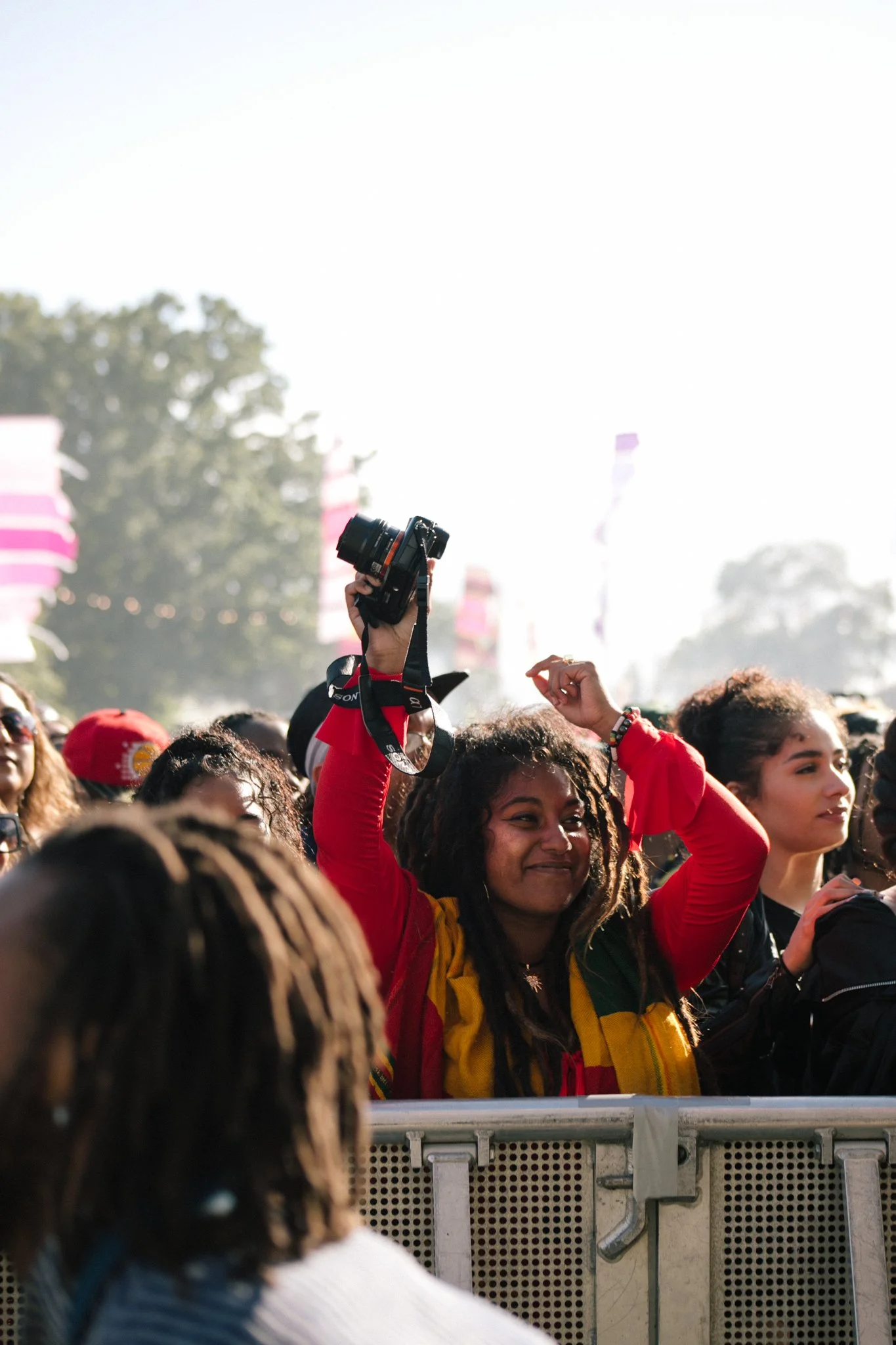 A diverse group of people at an outdoor event, with a woman in a red jacket raising her hand and holding a camera above the crowd. The scene has trees and flags in the background.