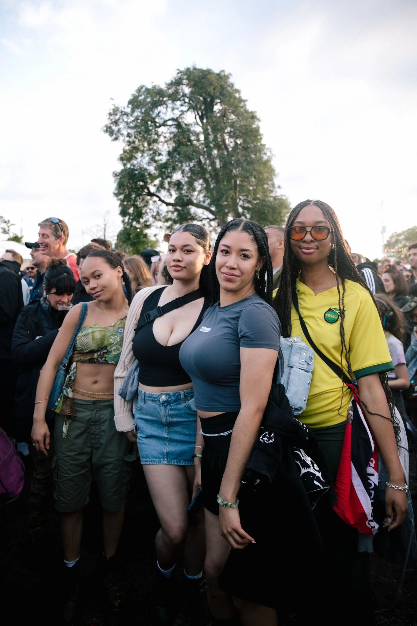 A group of young women posing together outdoors at a crowded event, with a large tree and a bright sky in the background.