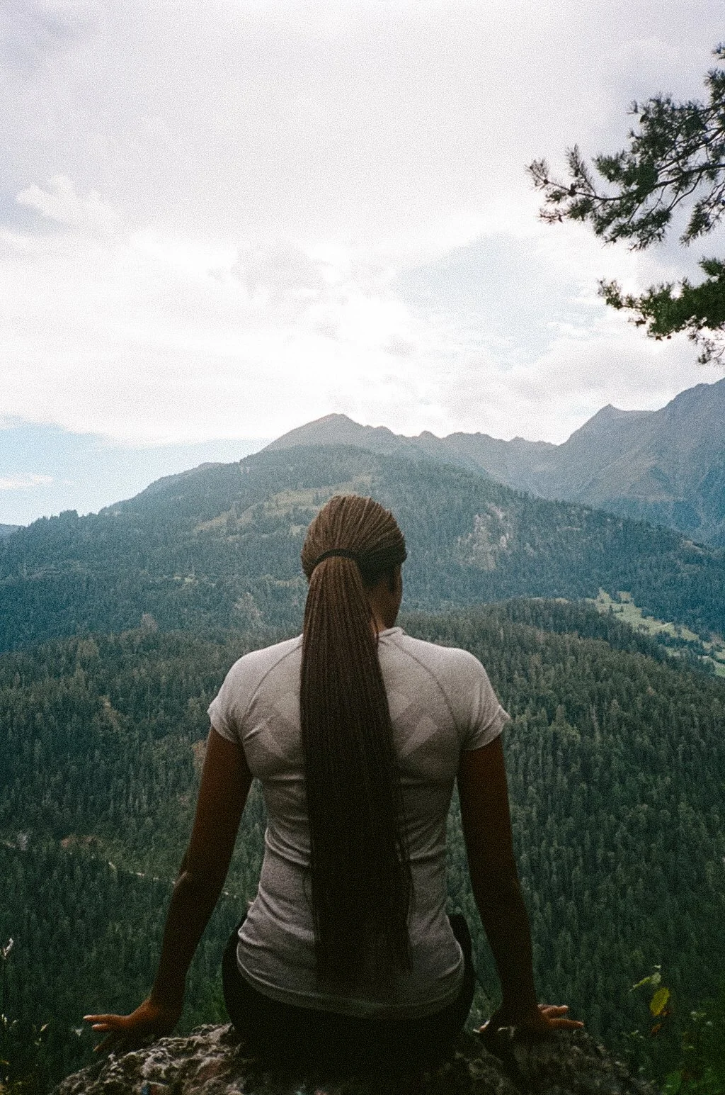 A woman with long braided hair sitting on a rock, overlooking a forested mountain landscape with mountains in the background and a cloudy sky.
