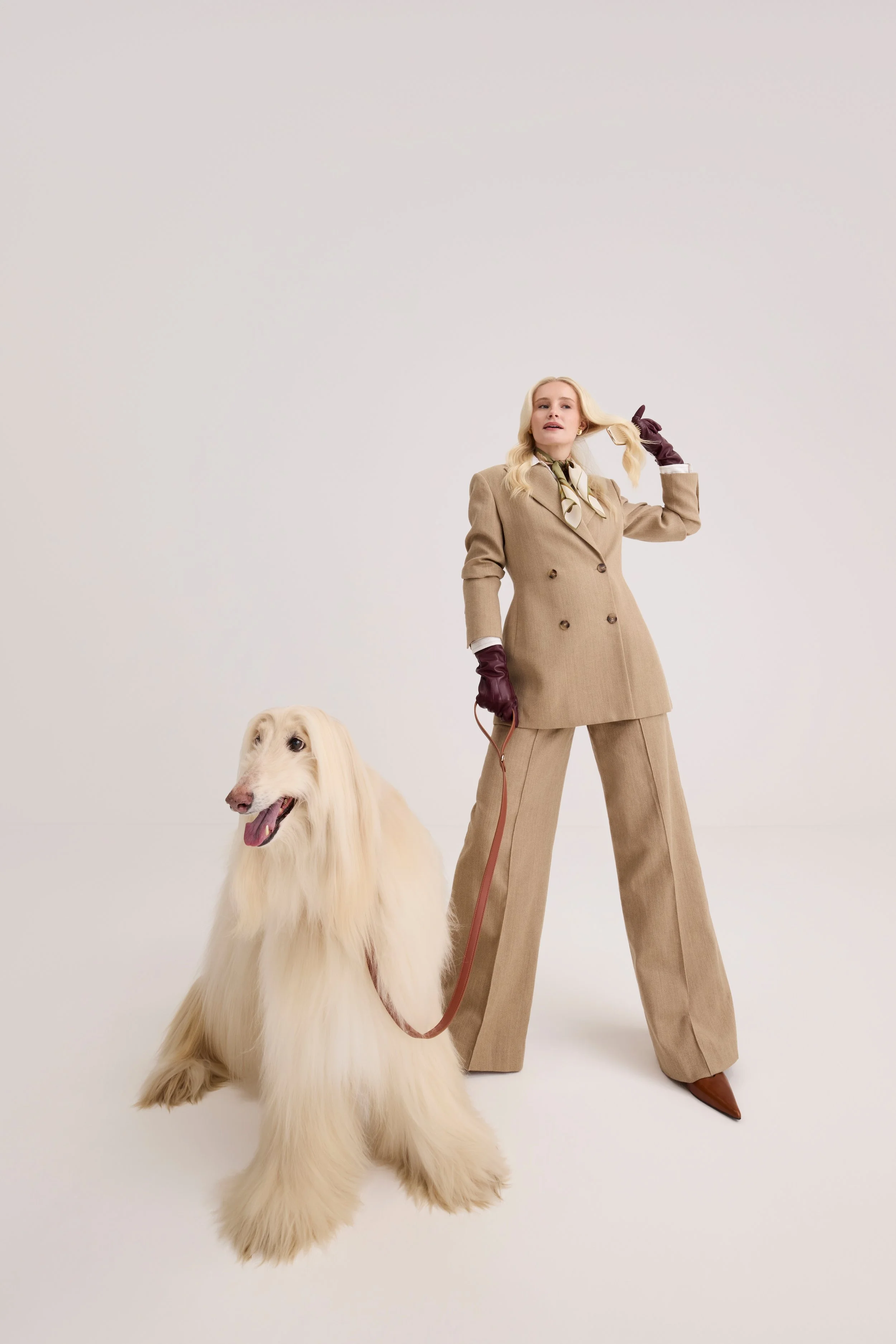 A woman with blonde hair in a beige suit, wearing dark gloves, standing on a white background, holding a leash attached to a large, fluffy, cream-colored dog with long hair and a happy expression.
