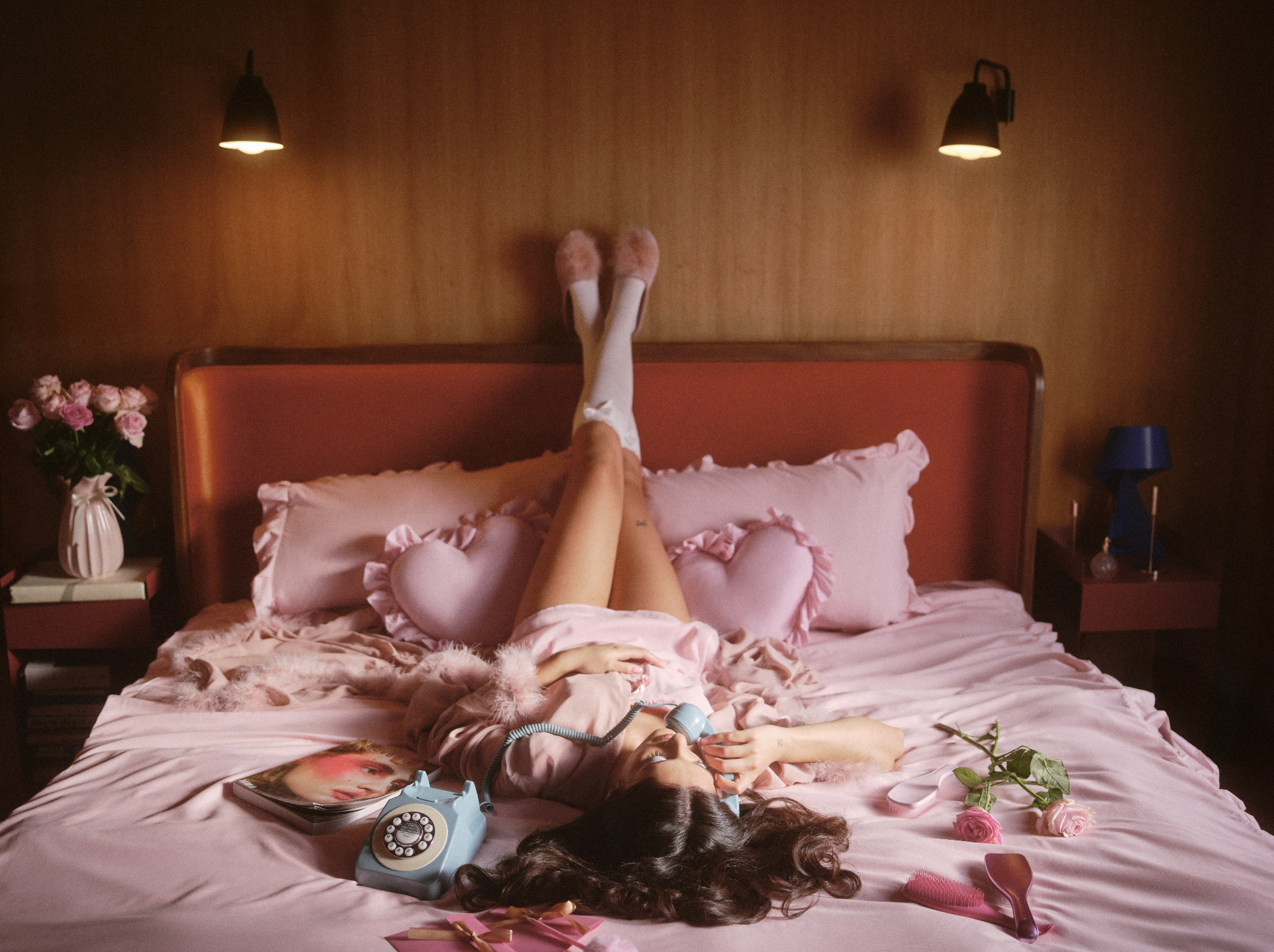 A person lying on a pink bed with pink ruffled pillowcases, wearing pink fluffy slippers and white stockings. Their legs are raised and resting against the headboard. The bedside tables hold flowers, a magazine, a vintage telephone, and various acces