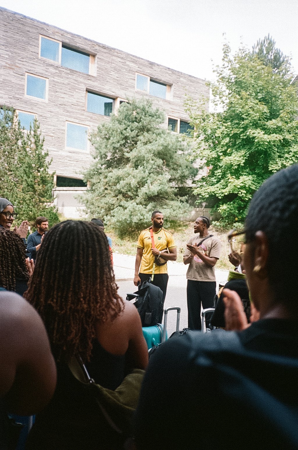 A group of people gathered outdoors, some with suitcases, listening to two men speaking to the crowd. One man wears a yellow shirt and holds a bag, while the other wears a beige shirt and claps. There are trees and a modern building in the background