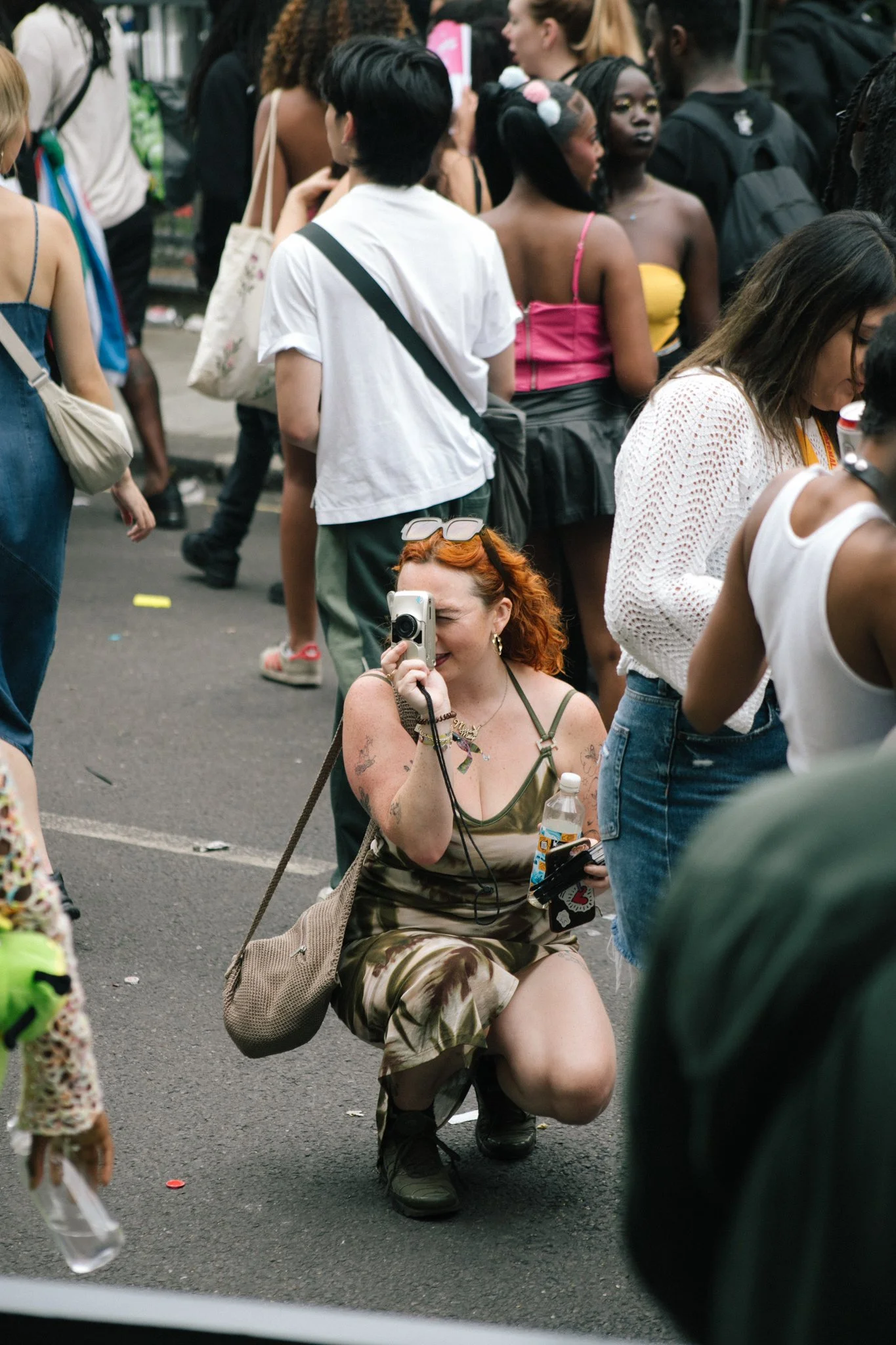 A woman with red curly hair taking a photo with a camera while kneeling on the ground at a crowded outdoor event. She is wearing a camouflage dress and has tattoos on her arms.