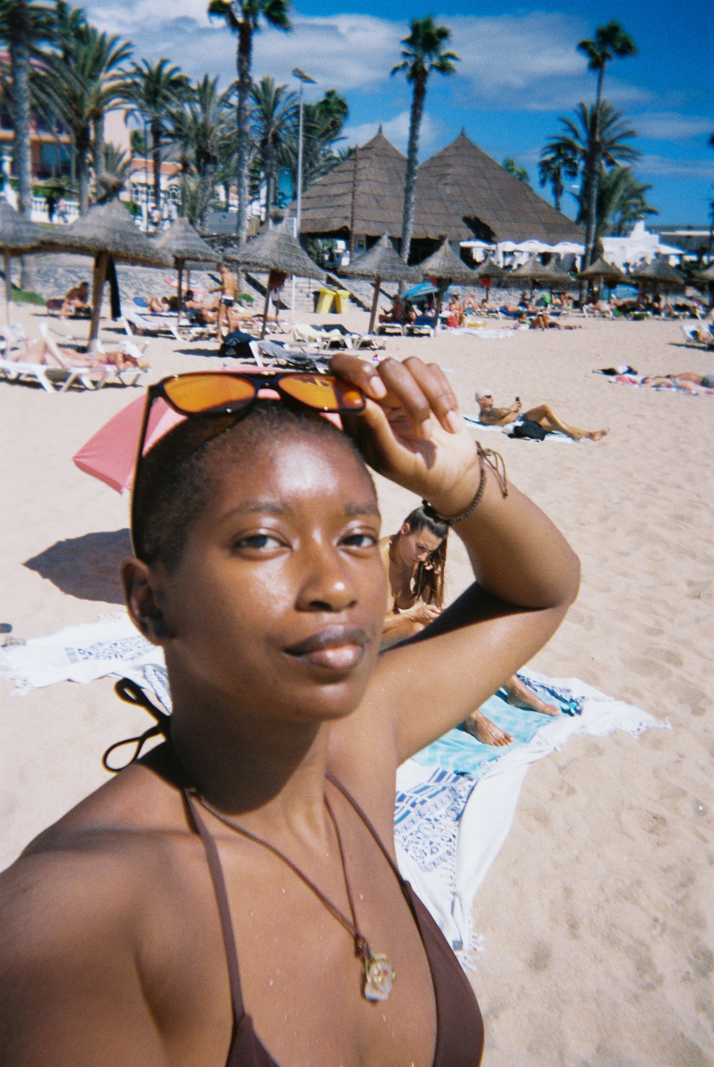 A woman at the beach taking a selfie, with beach umbrellas, sunbathers, palm trees, and thatched roof structures in the background.