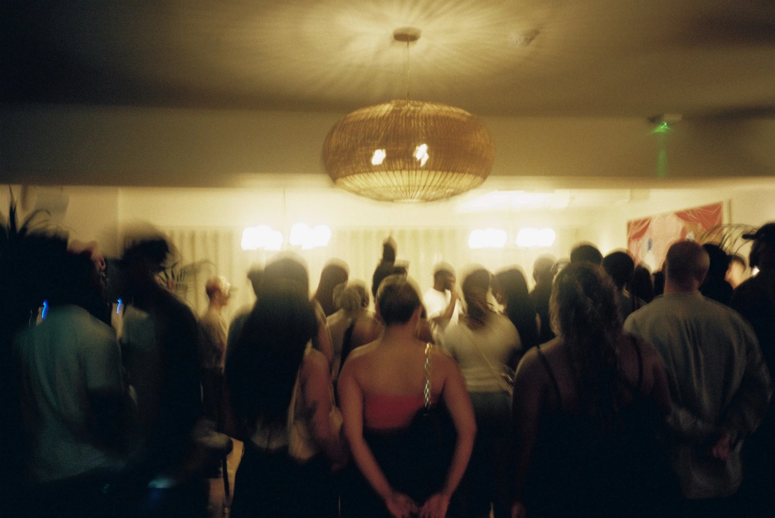 A crowded gathering with people listening to live music under warm lighting. There is a large, round, woven pendant light hanging from the ceiling.