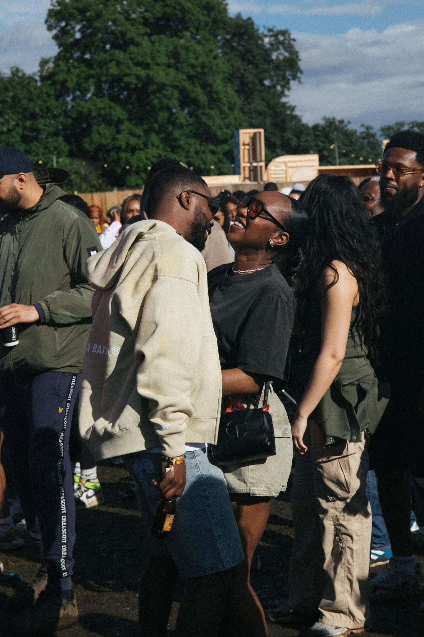A group of people outdoors at a social gathering, with some individuals smiling and engaging with each other. The background shows green trees and a partly cloudy sky.