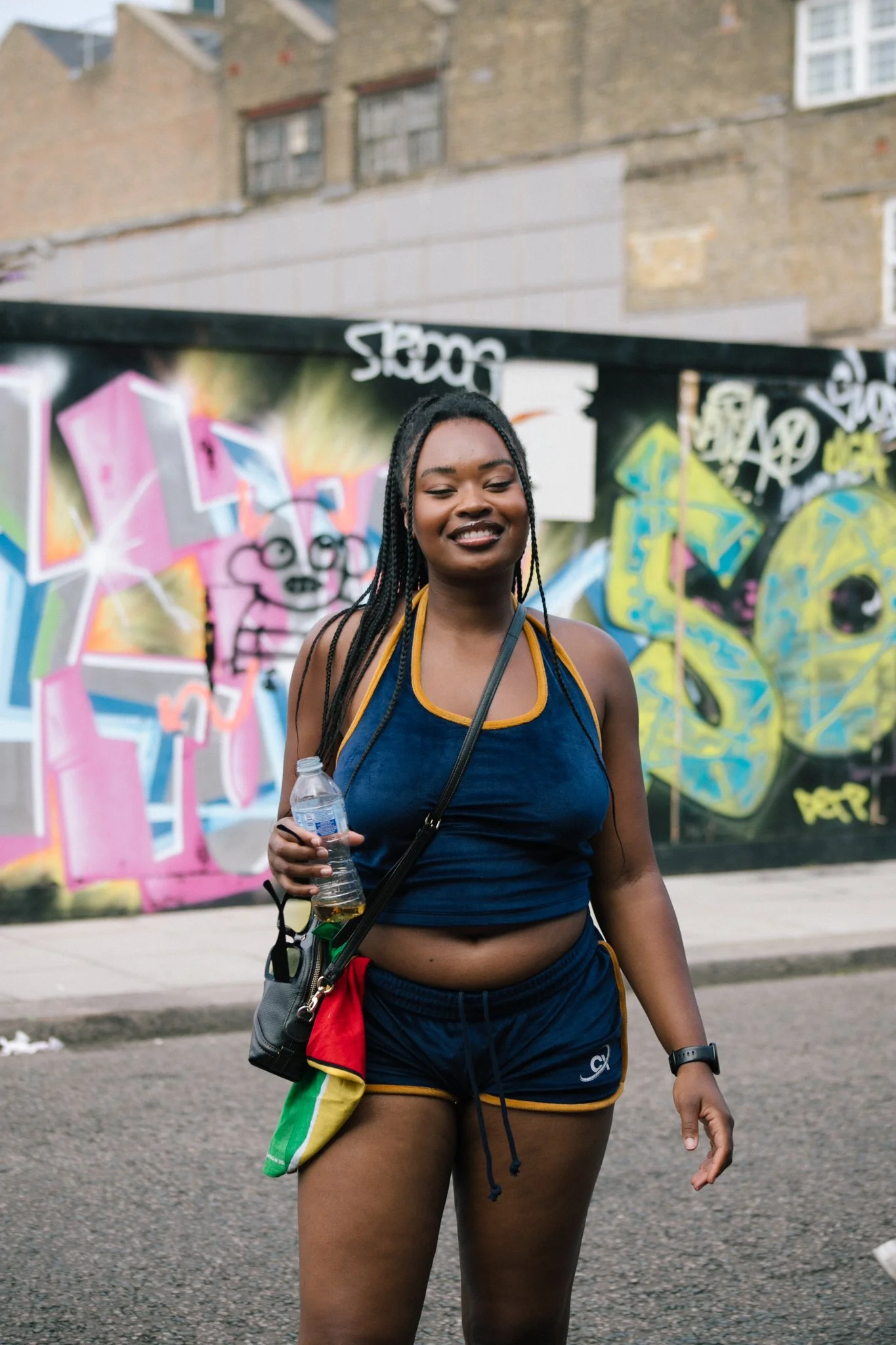 A young woman with braided hair smiling and holding a water bottle, standing on a street with graffiti-covered wall in the background.