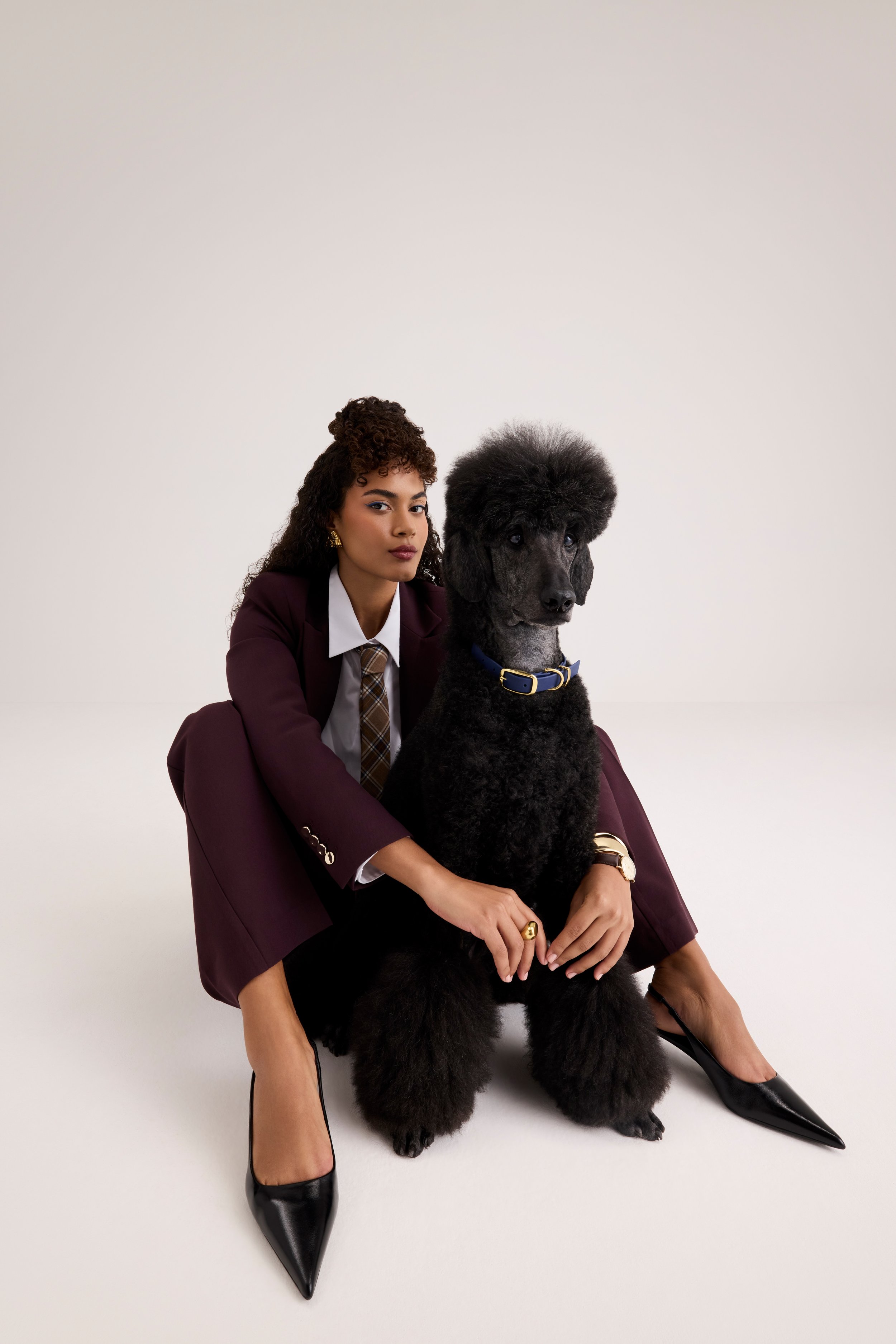 A woman in a maroon suit with a white shirt and plaid tie, sitting with her arm around a large black poodle, both on a plain white background.