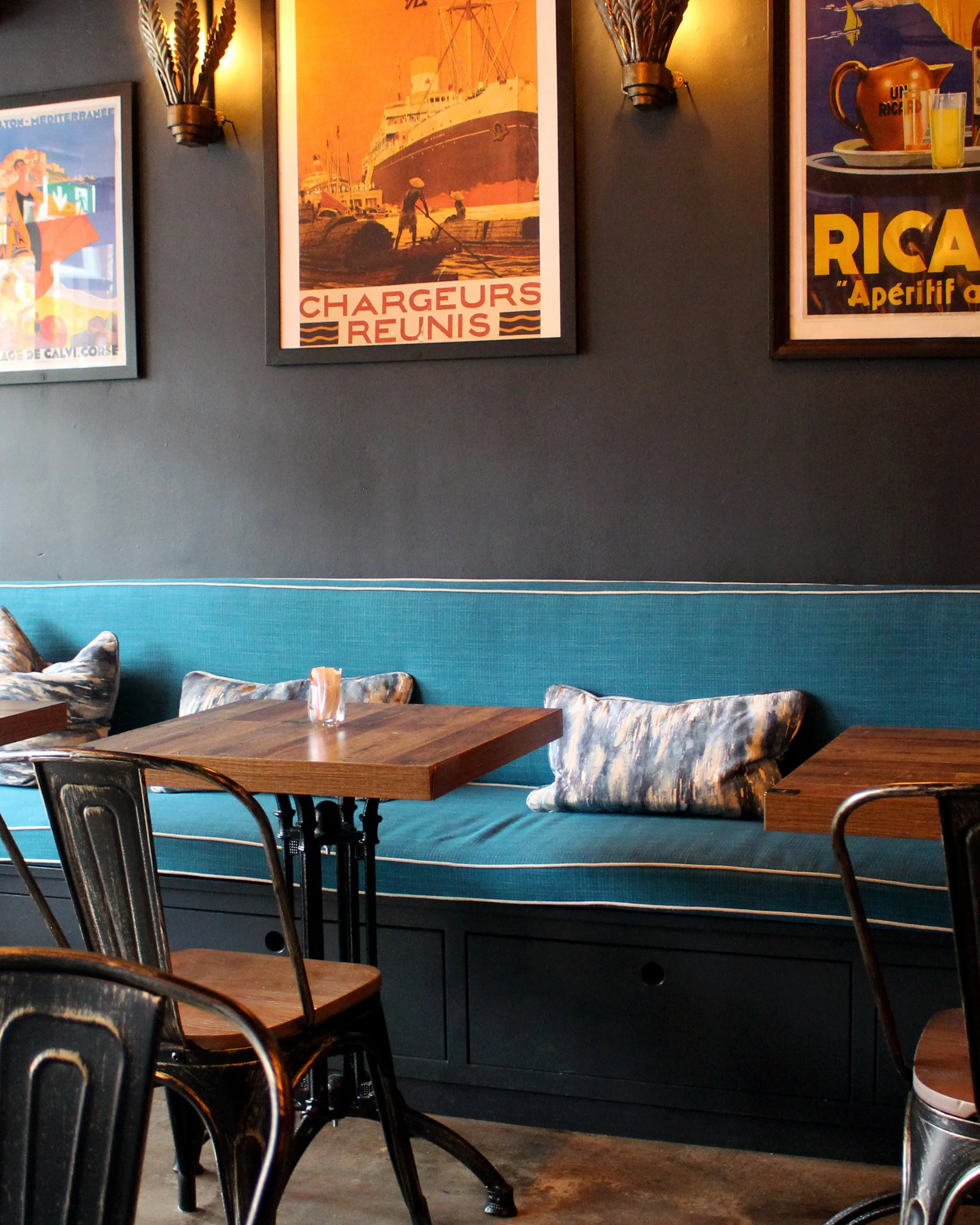 Seating area inside Sev7en café in Bray with a blue banquette, wooden tables, black chairs, and framed vintage-style wall posters.