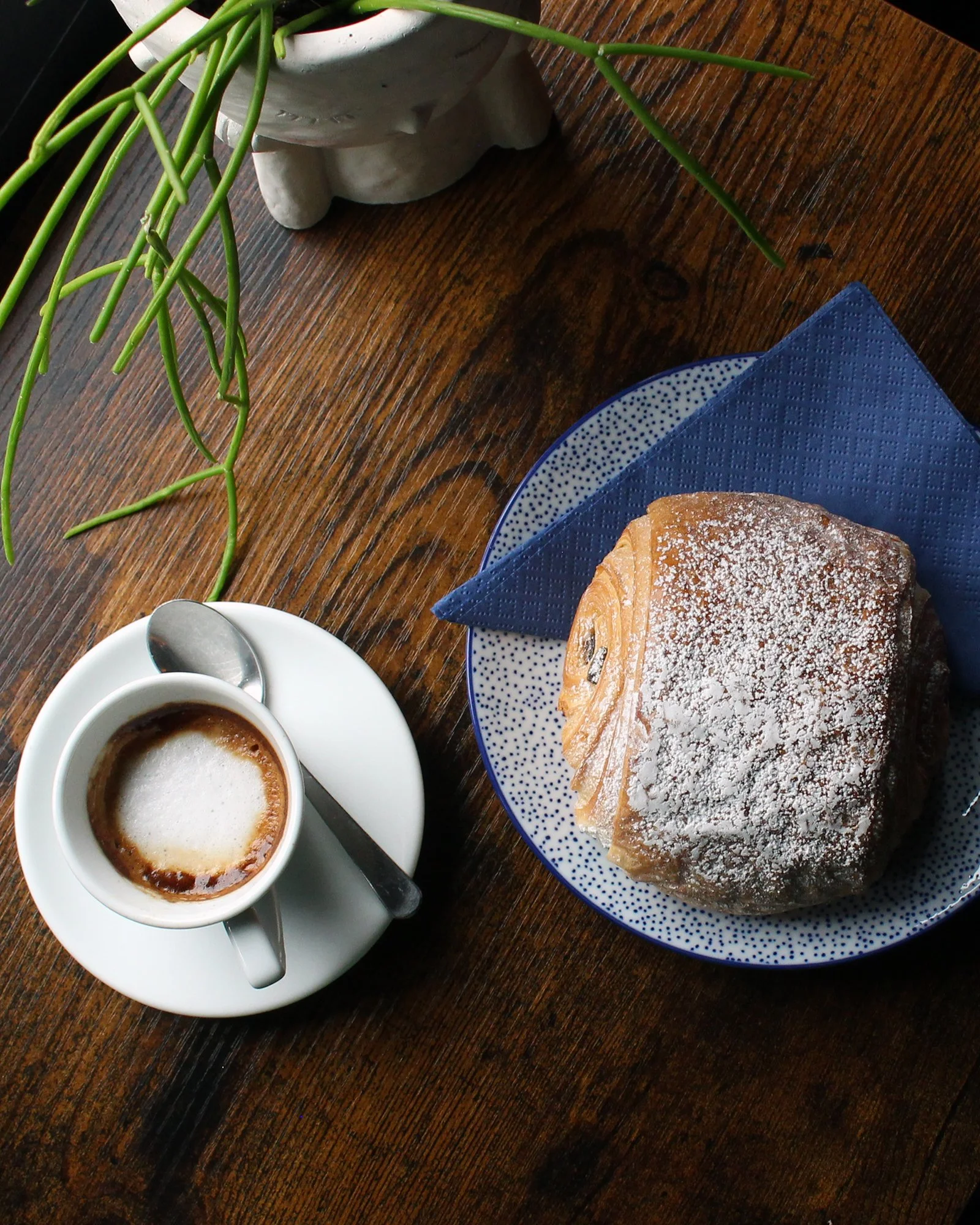 Fresh macchiato served with a powdered sugar pain au chocolat on a wooden table at SEV7EN Cafe, styled with a plant and natural light.