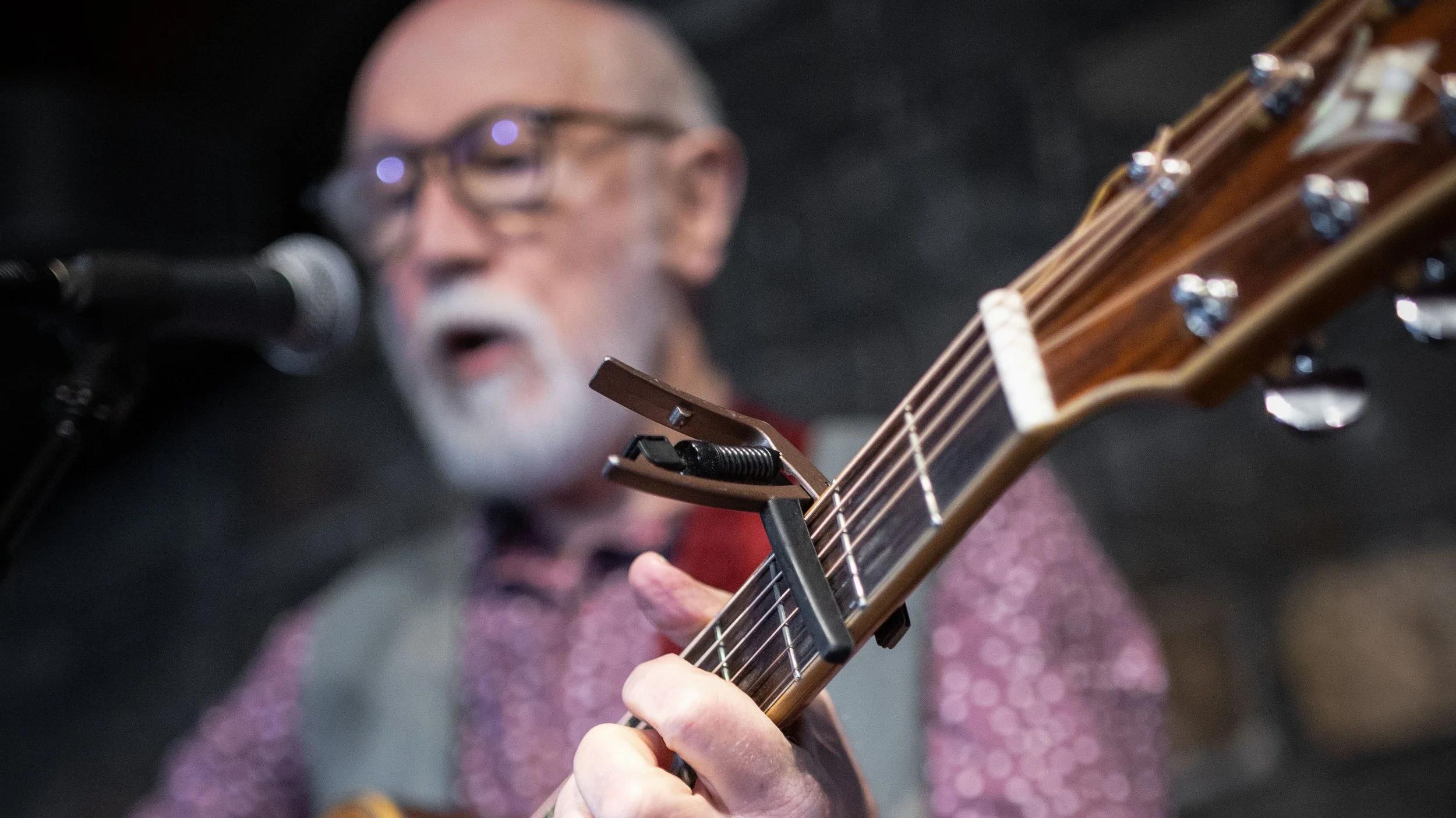 A blurred close-up of a Alan Reid, a Scottish folksinger, with a guitar and capo, with a microphone in front